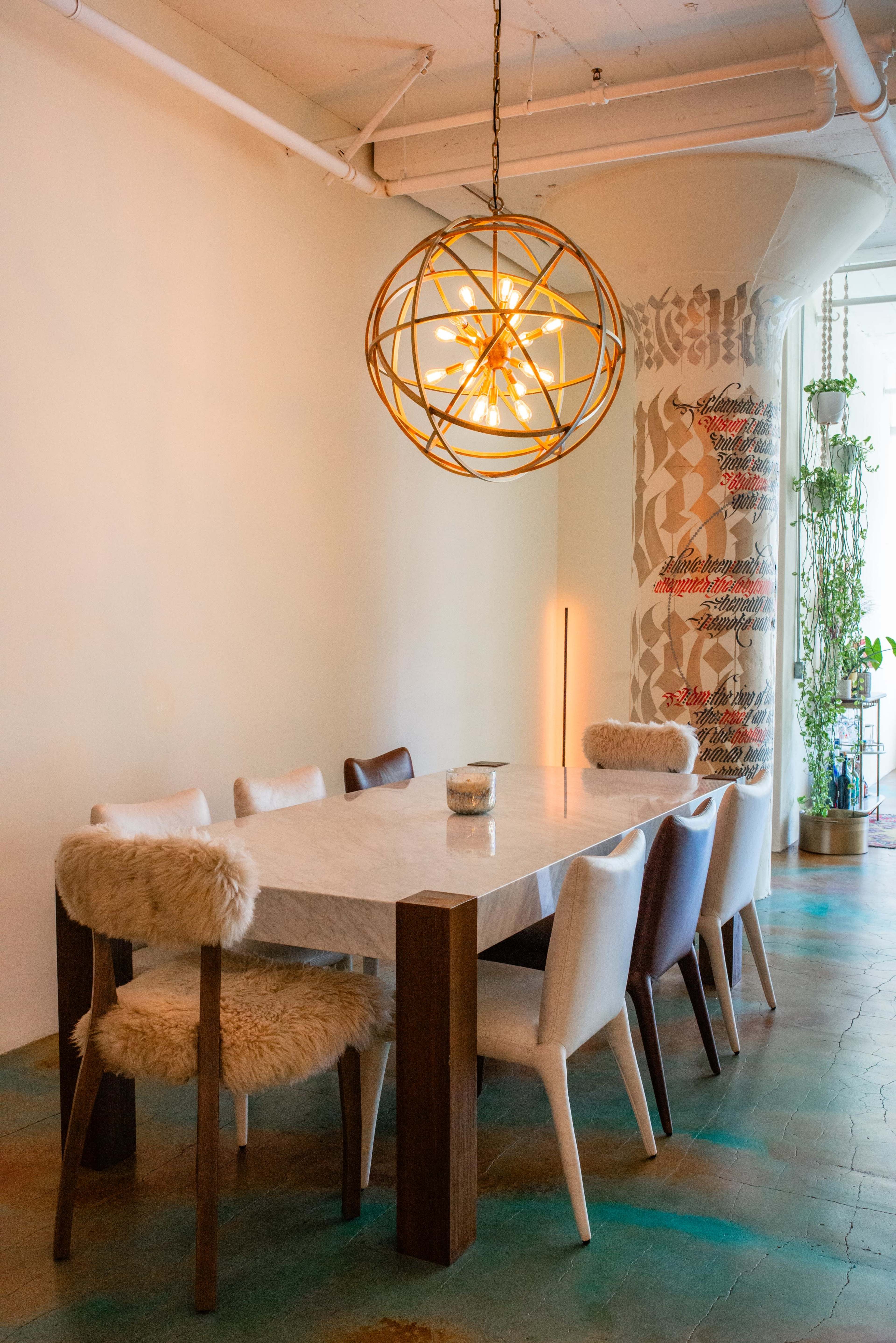 A spacious dining room features a long table surrounded by chairs in various colors, illuminated by an ornate pendant light.