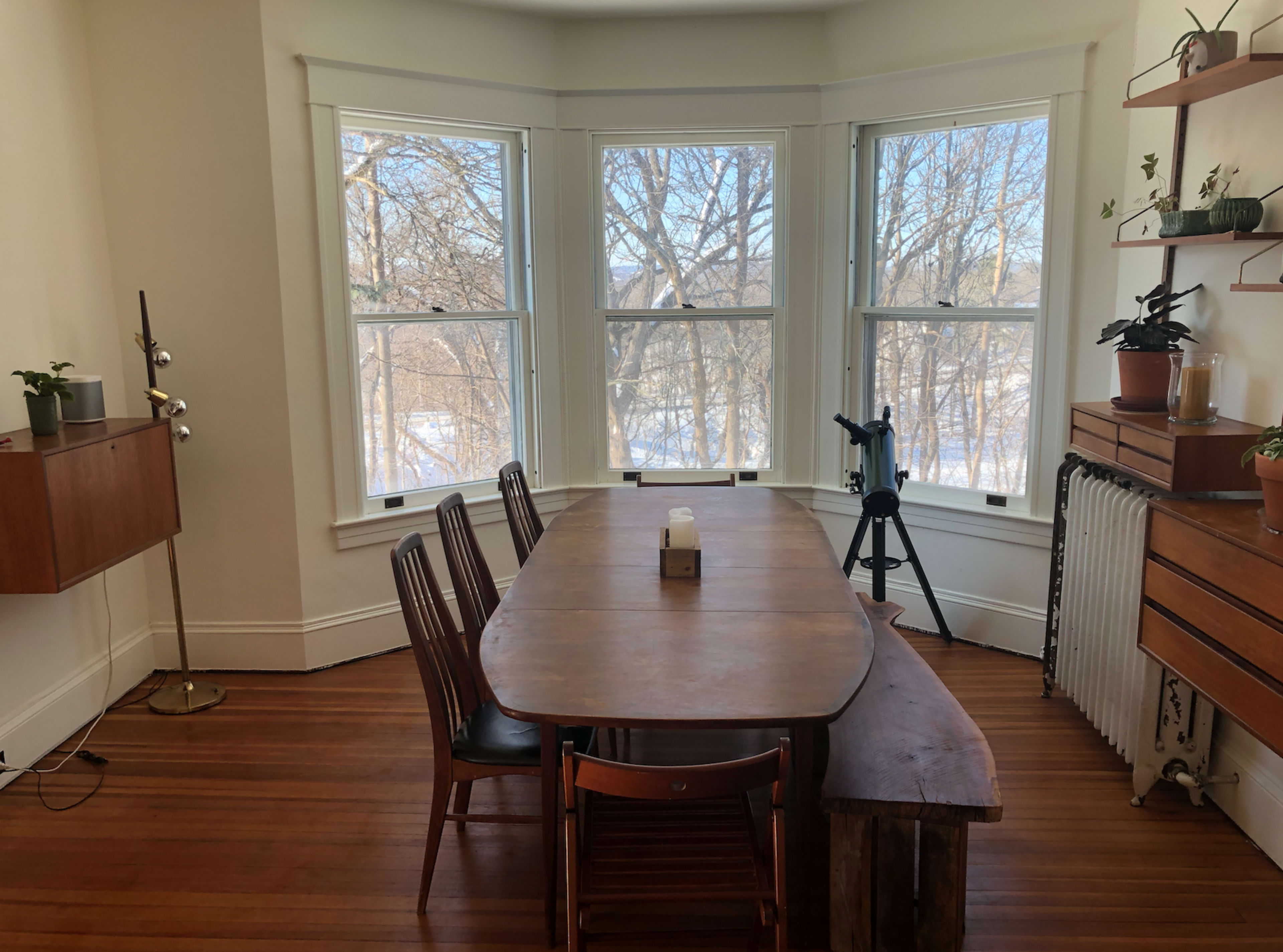 A wooden dining table with several chairs is positioned in a sunlit room featuring bay windows, with trees visible outside.