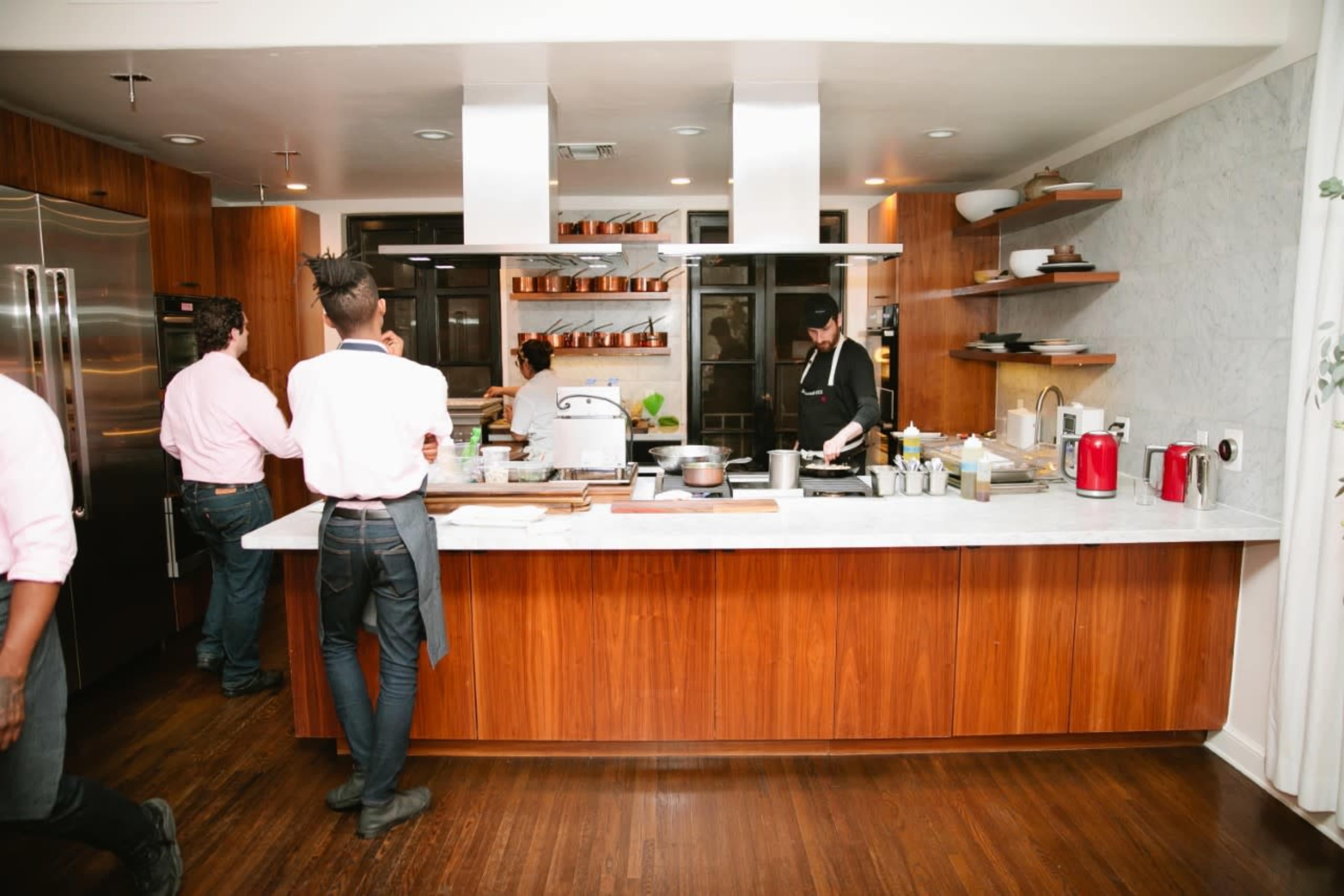 A modern kitchen features a long countertop with various cooking utensils and appliances, while several individuals in casual attire work on food preparation.