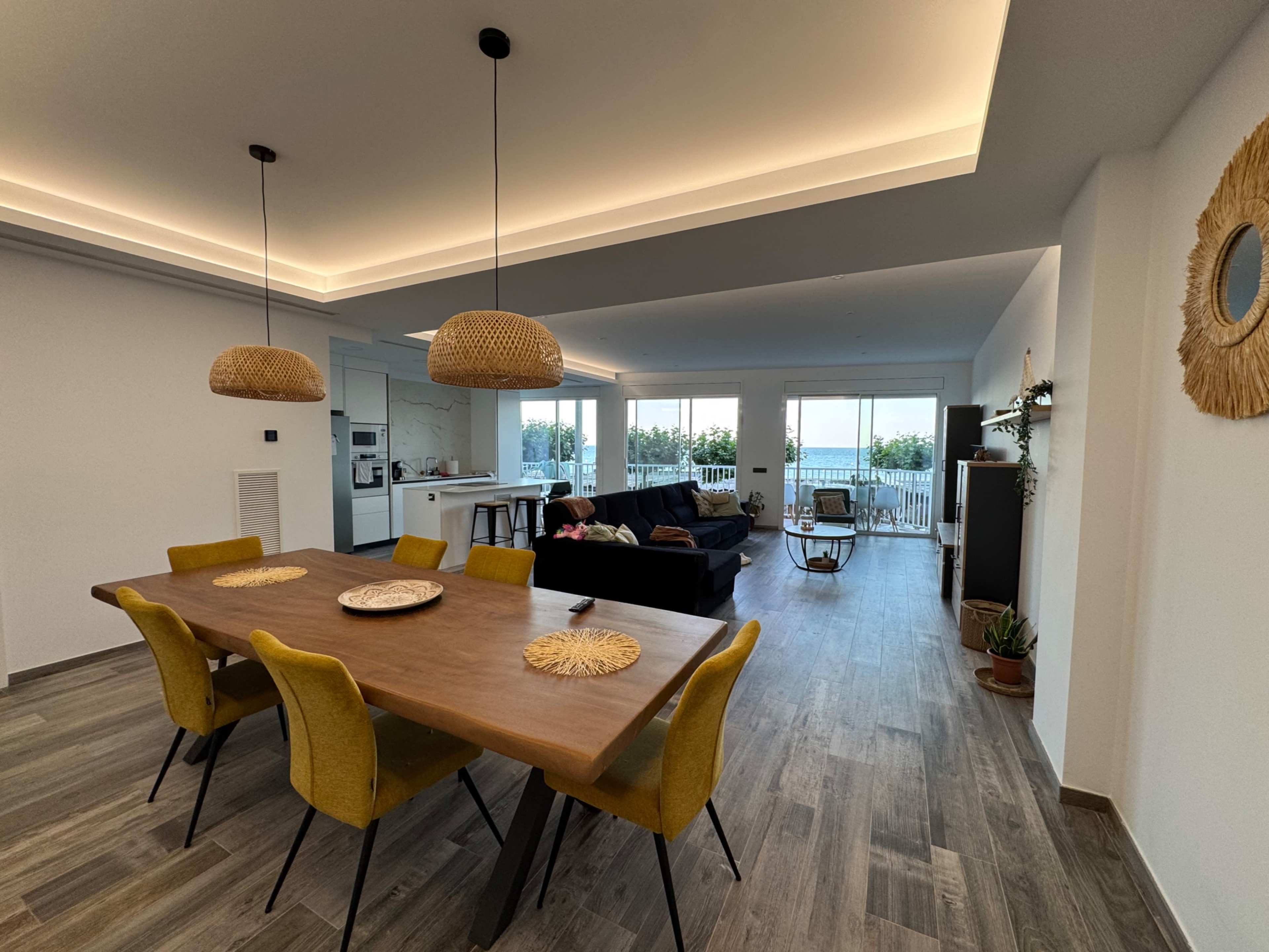 The image shows a modern open-concept dining and living area featuring a wooden dining table with yellow chairs, a dark sofa, and large windows that provide natural light and a view of the outdoors.
