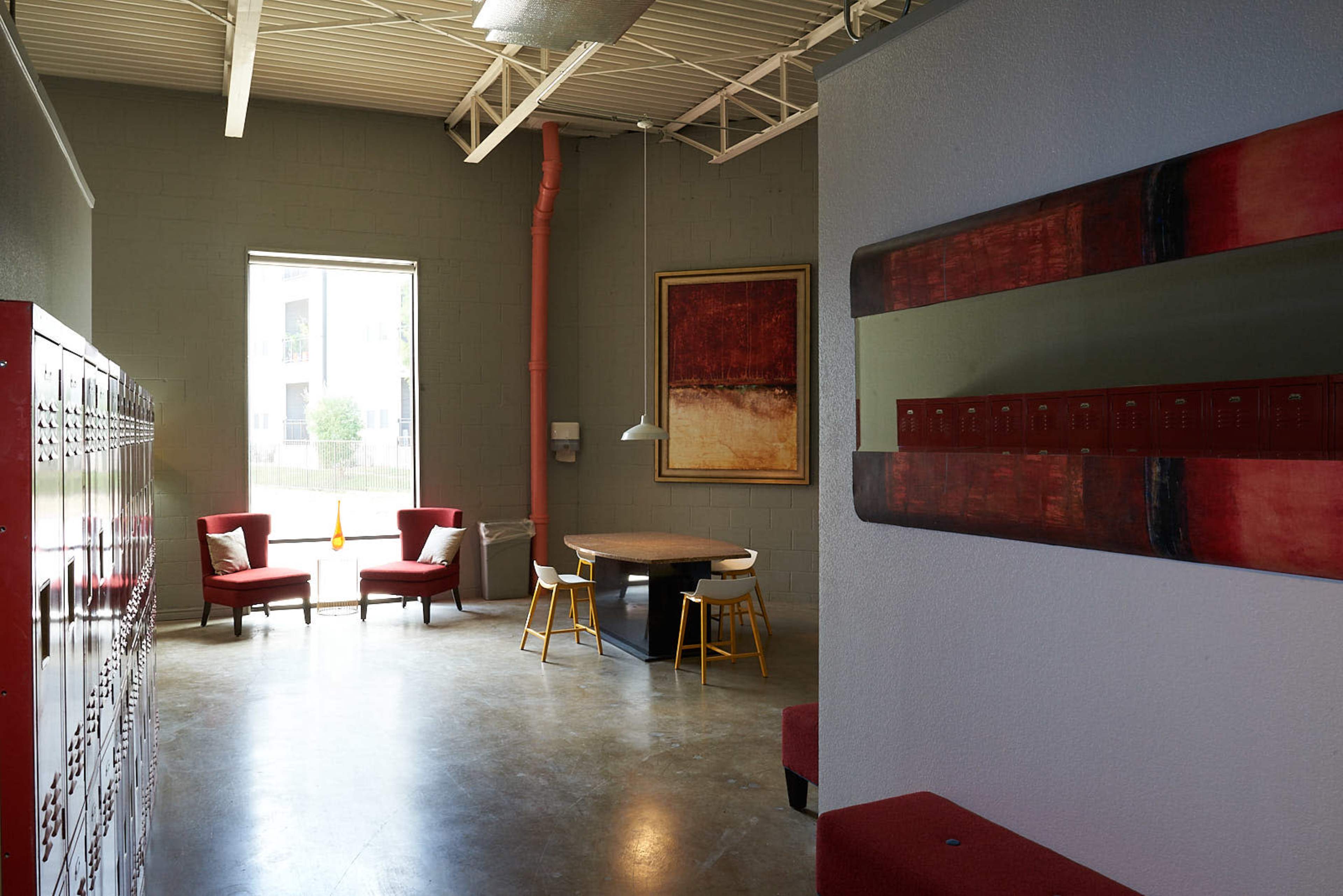 A modern, open space with red lockers lining one wall, a round table with chairs in the center, and two red armchairs near a large window.