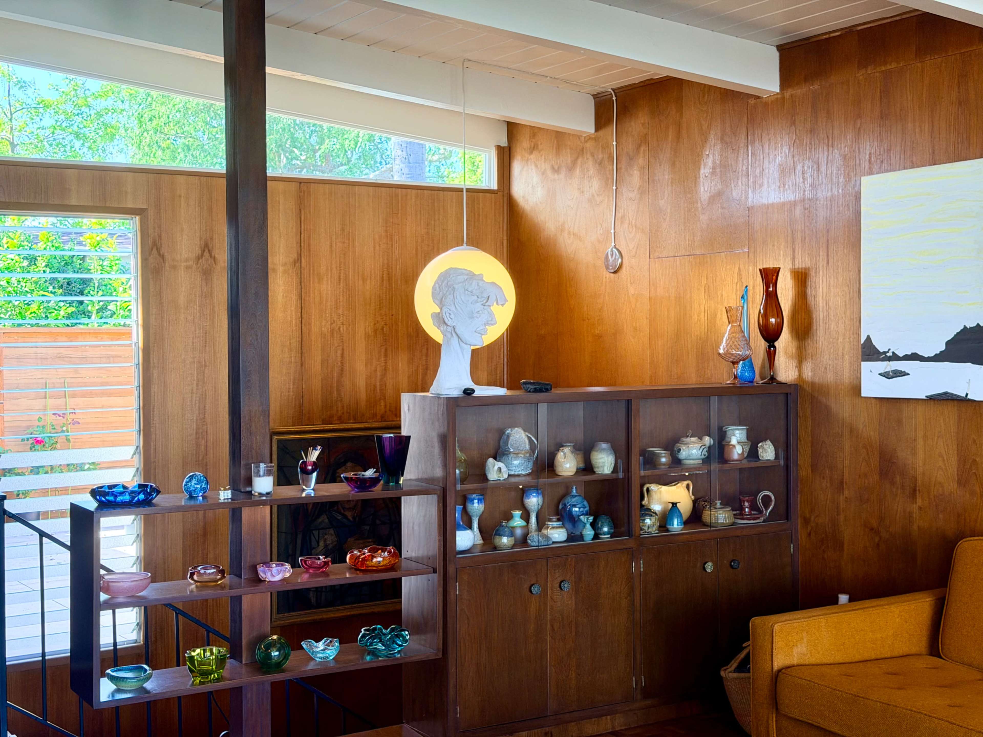 The image shows a mid-century modern living room with wooden walls, a display cabinet filled with colorful glassware, and a cozy orange sofa.