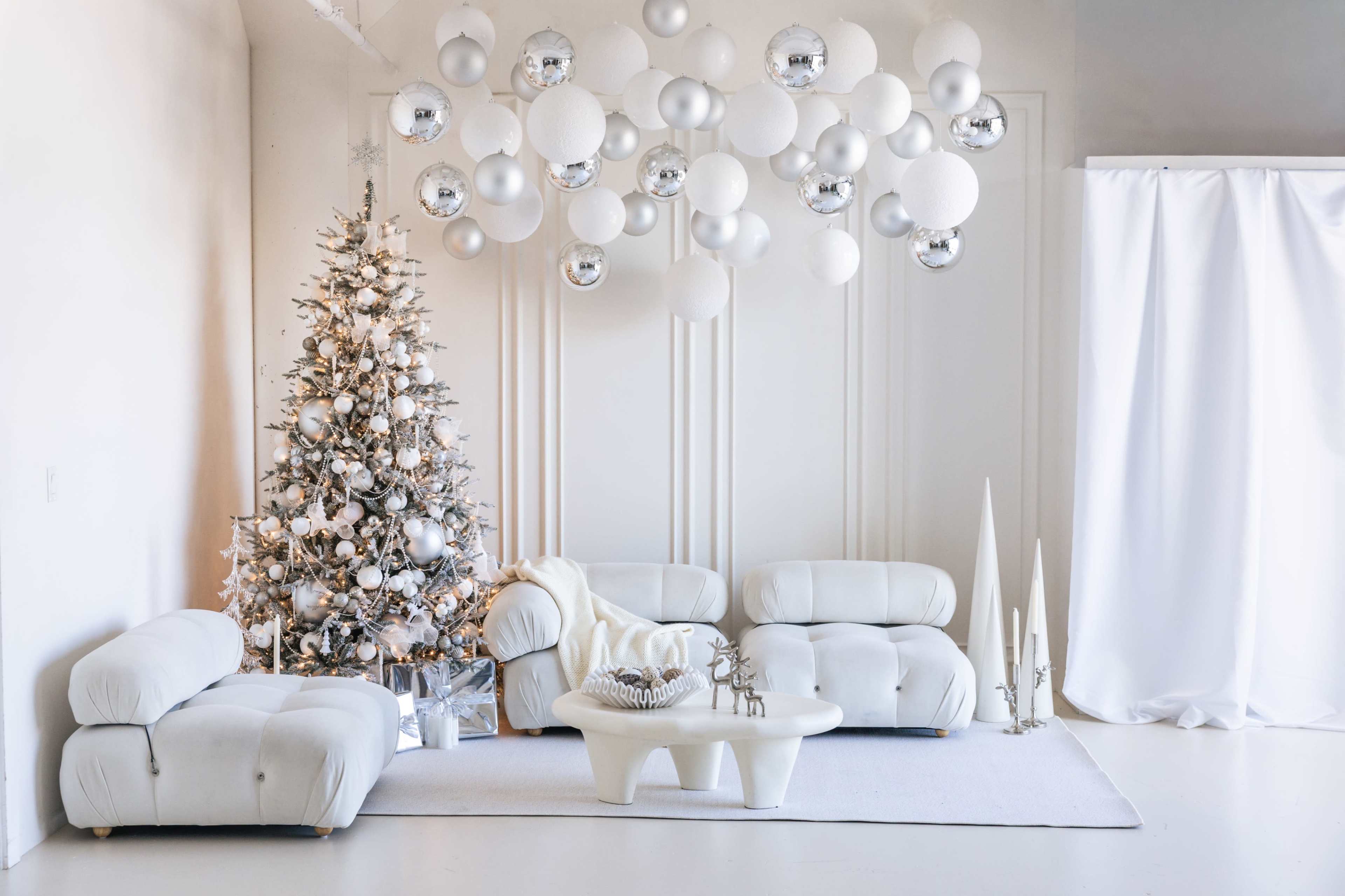A decorated Christmas tree stands next to modern white furniture in a minimalist room with circular ornaments hanging from the ceiling.