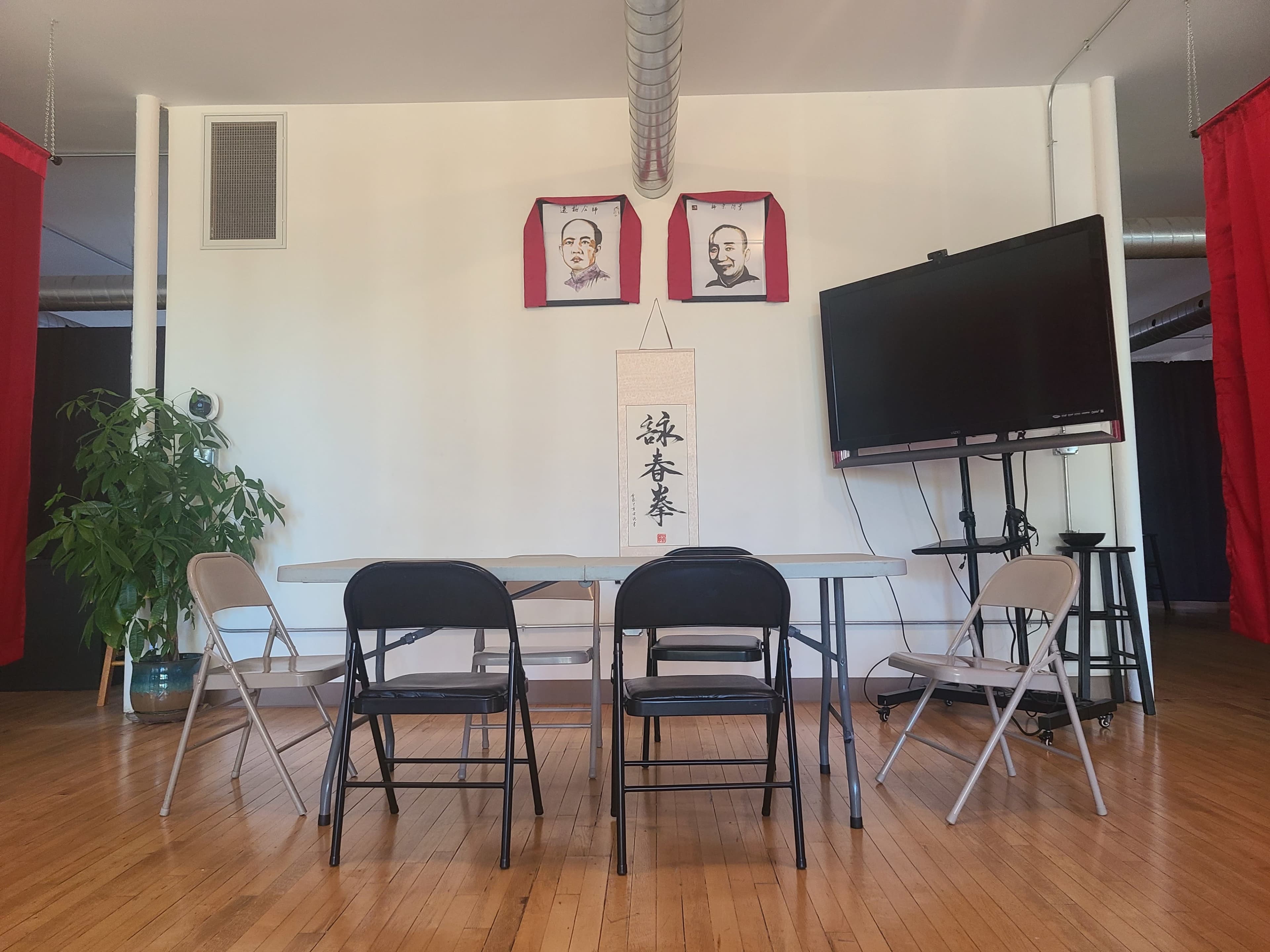 A meeting room features a rectangular table surrounded by black and white folding chairs, with two portraits on the wall and a large screen on the right.