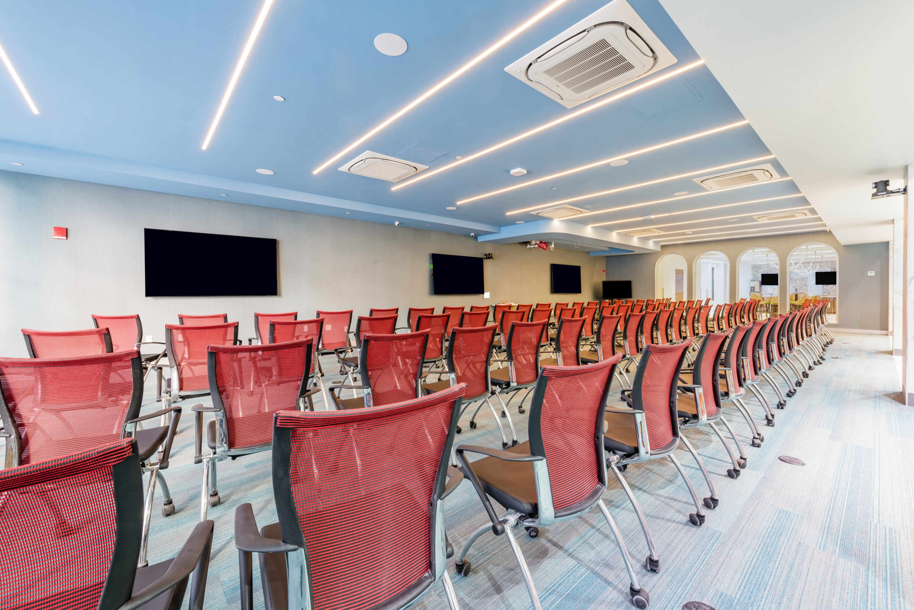The image shows a modern conference room with rows of red mesh chairs facing two wall-mounted screens.