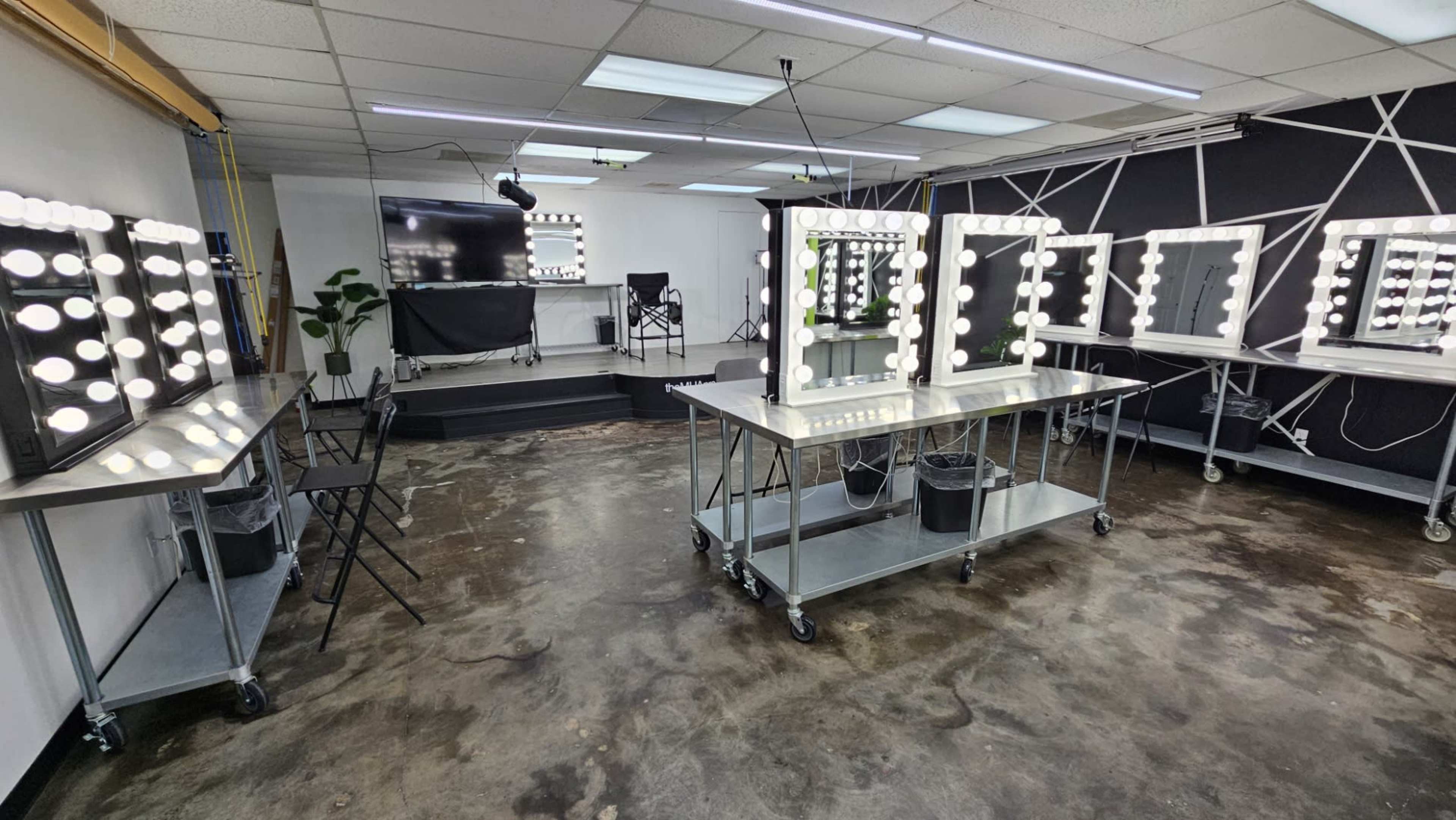 The image shows a makeup studio with several lighted mirrors on mobile tables, a black chair, and a television in the background.