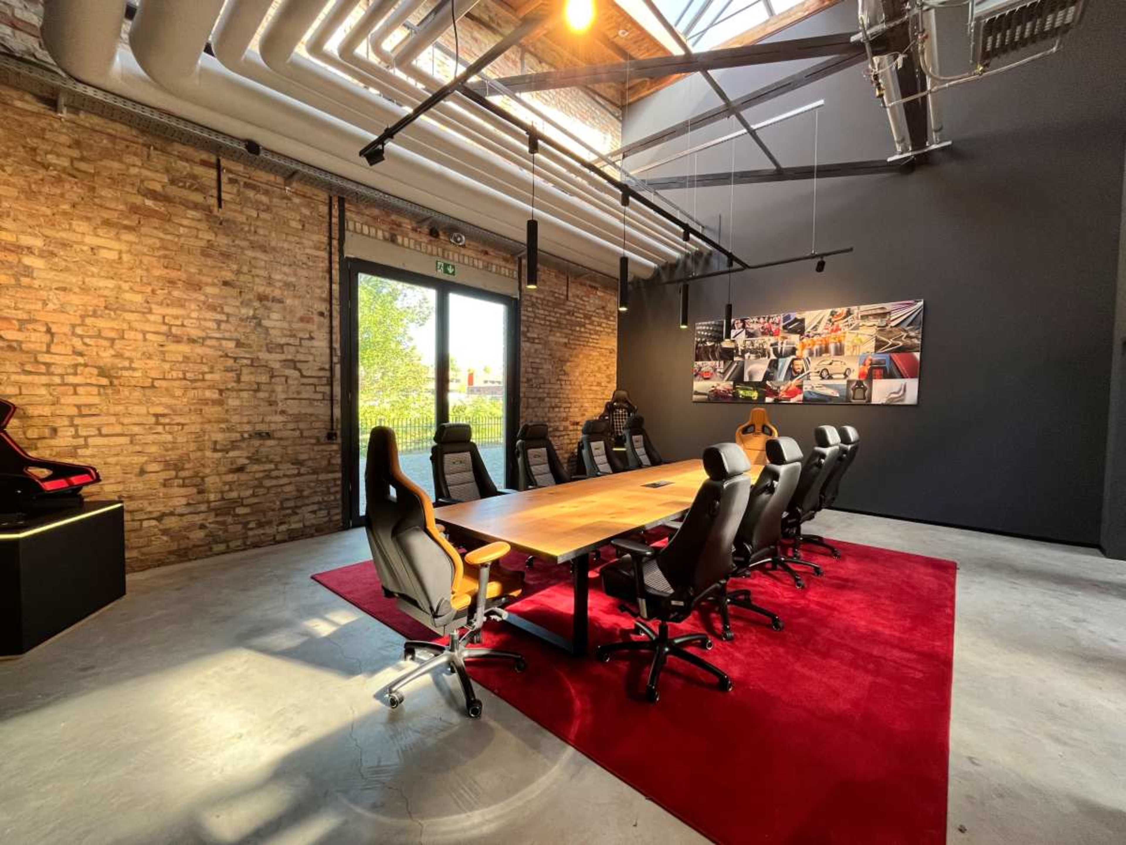 A modern conference room features a large wooden table surrounded by black and orange office chairs, with exposed brick walls and a large window letting in natural light.