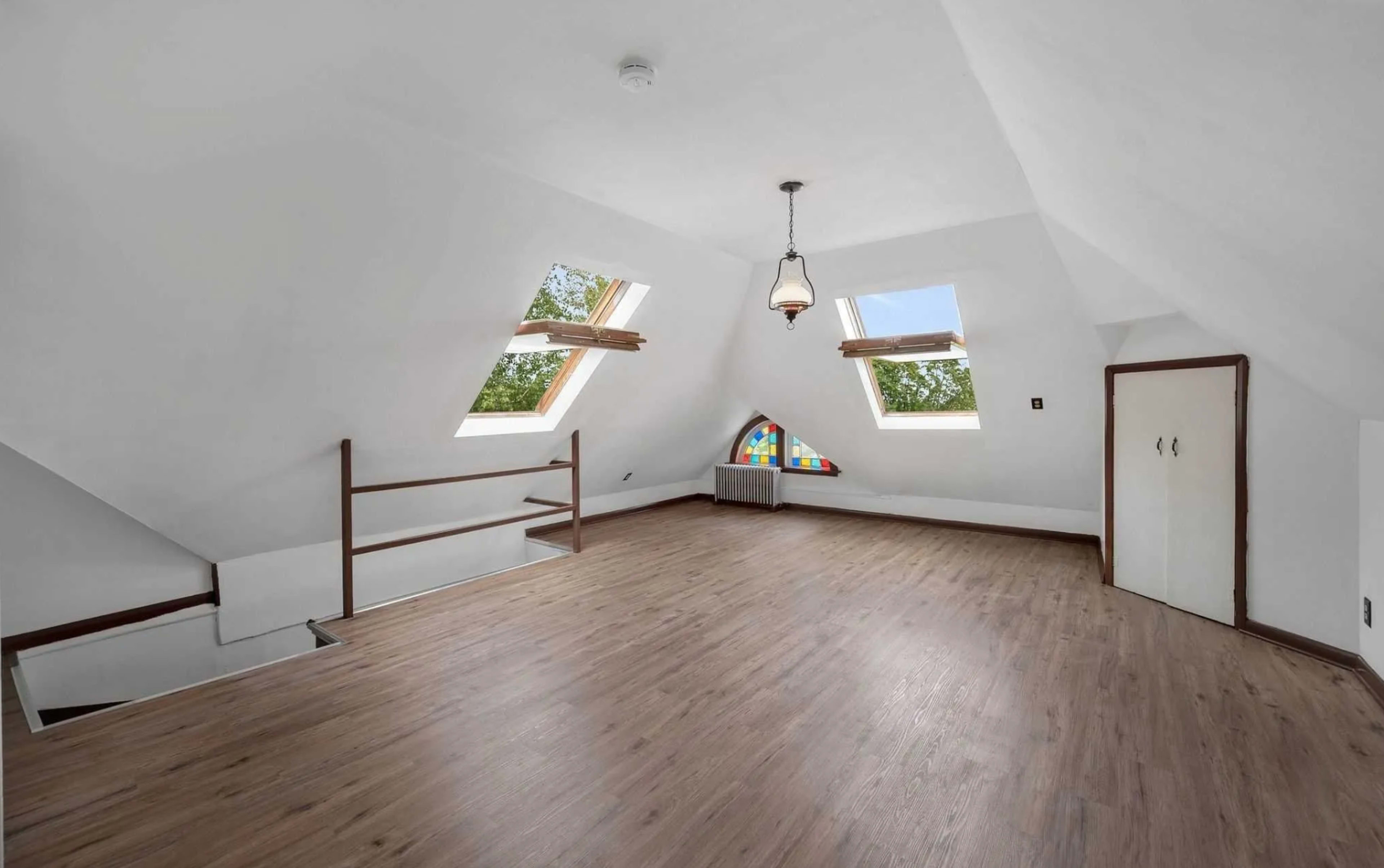 The image shows an empty attic room with sloped ceilings, two skylight windows, and a small stained glass window.
