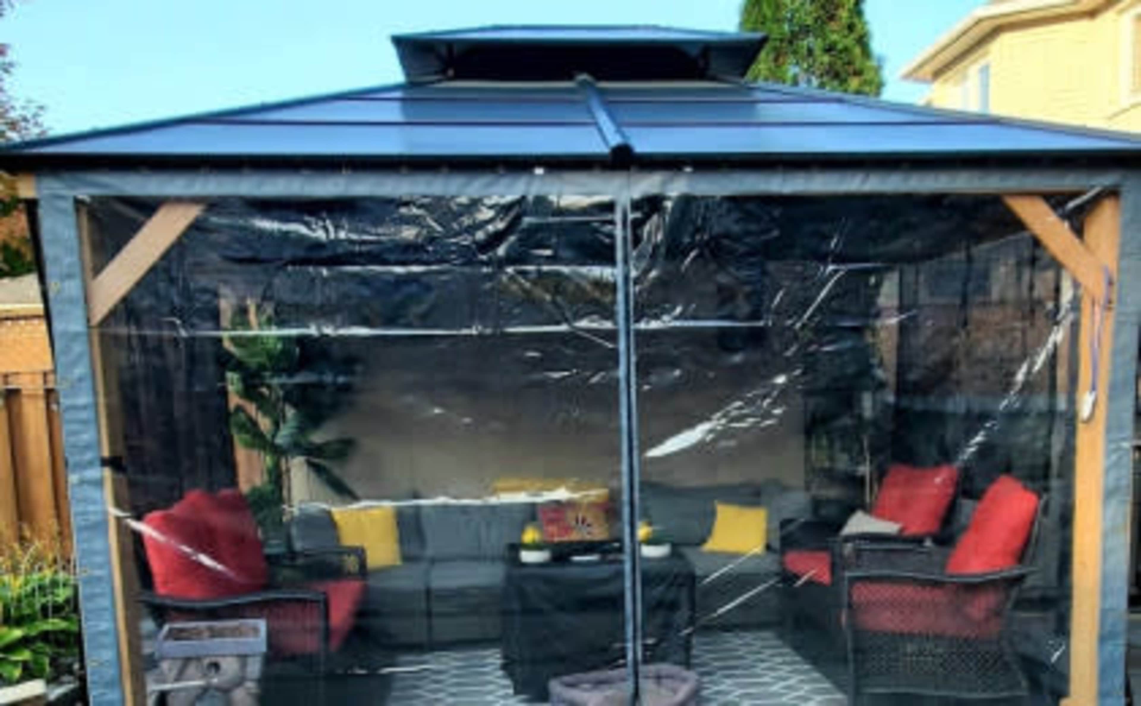 A gazebo with transparent side panels, containing a seating area with red and yellow cushions.