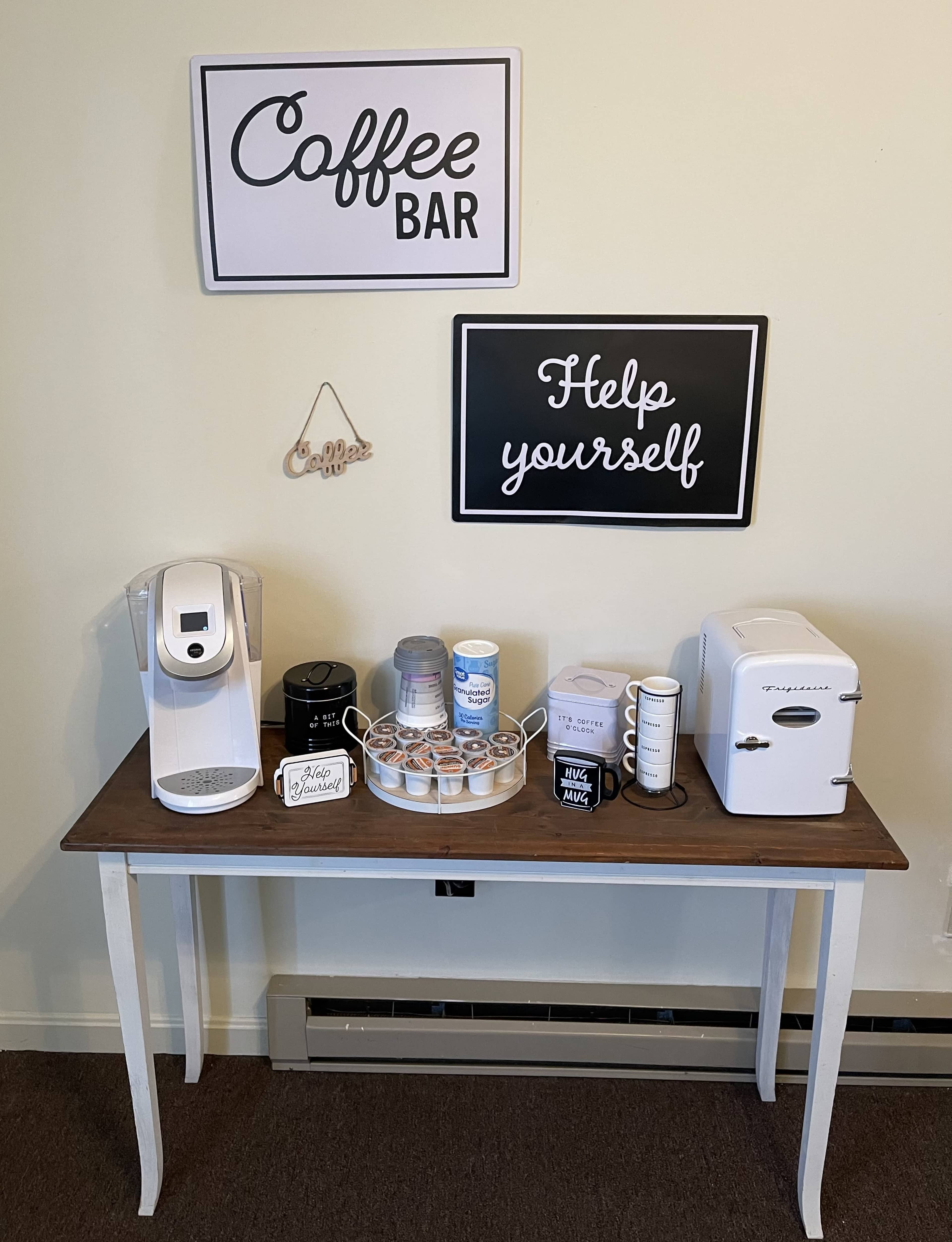 A coffee bar setup on a table, featuring a coffee maker, various coffee supplies, and decorative signs on the wall.