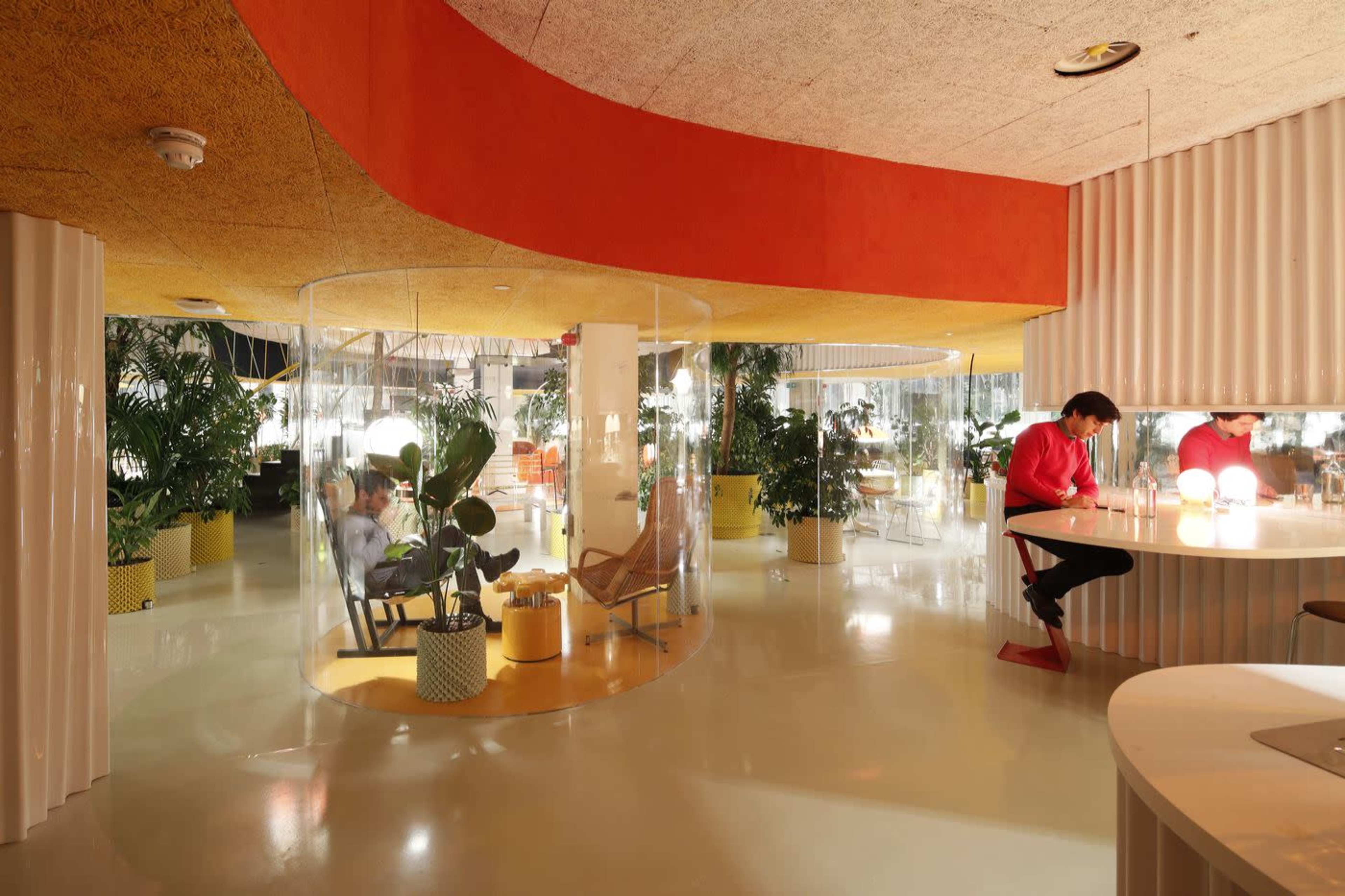 A modern interior featuring curved glass enclosures, vibrant orange and yellow accents, and indoor plants, with people engaging in various activities.