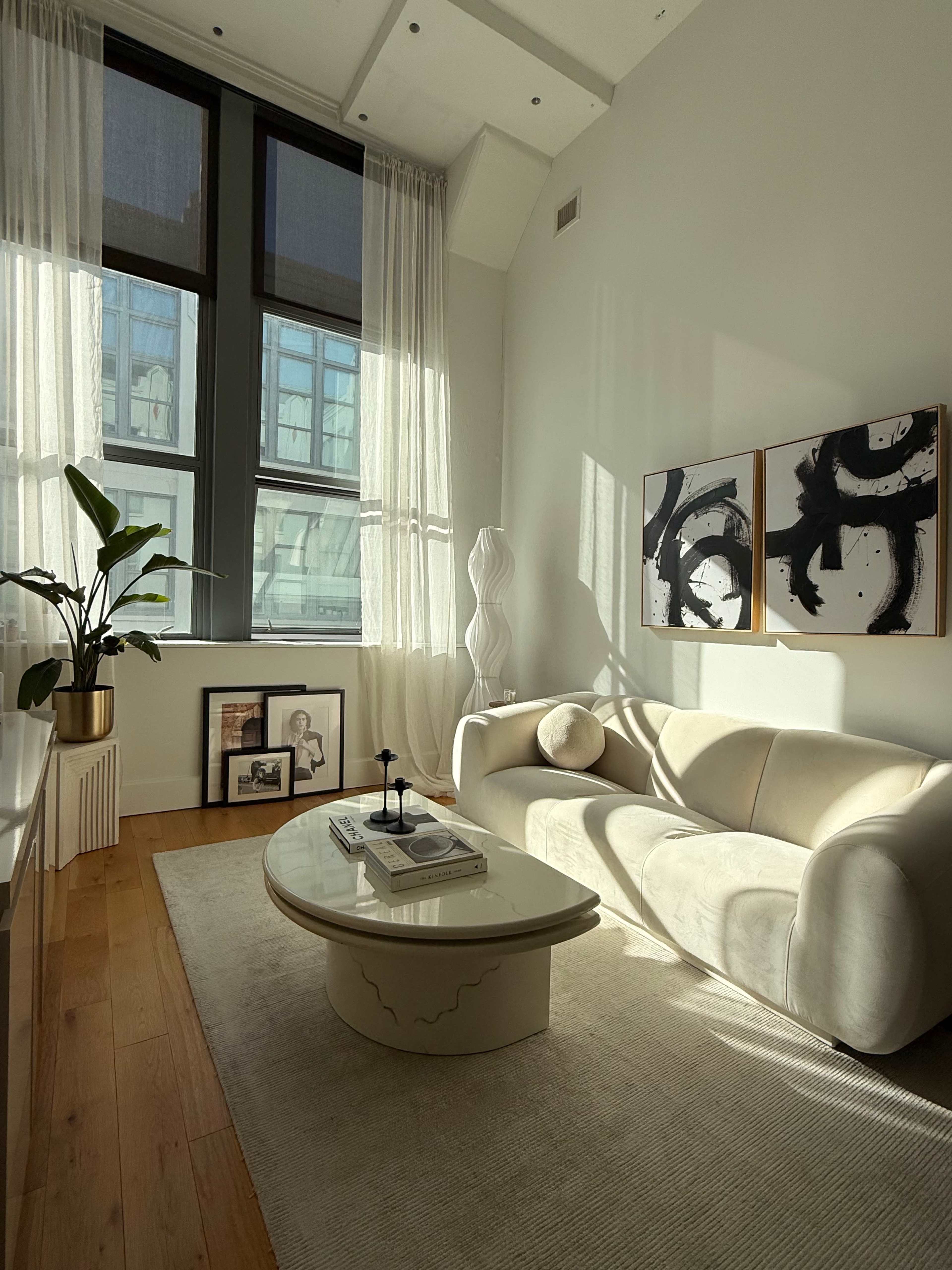 A modern living room features a light-colored sofa, a round coffee table, and large windows with sheer curtains, allowing sunlight to illuminate the space.