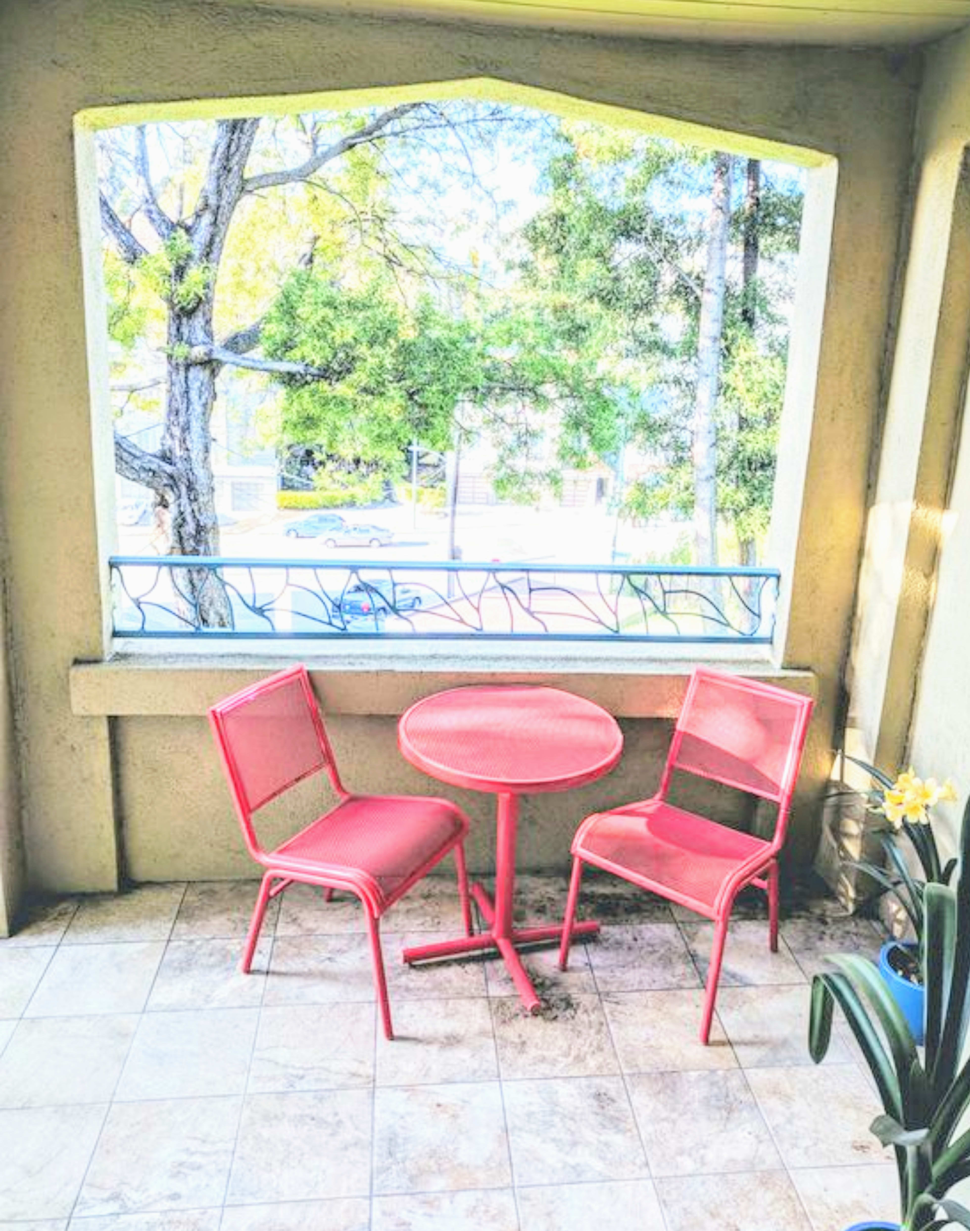 A small balcony features a round table and two red chairs beside a window view of trees and a street.