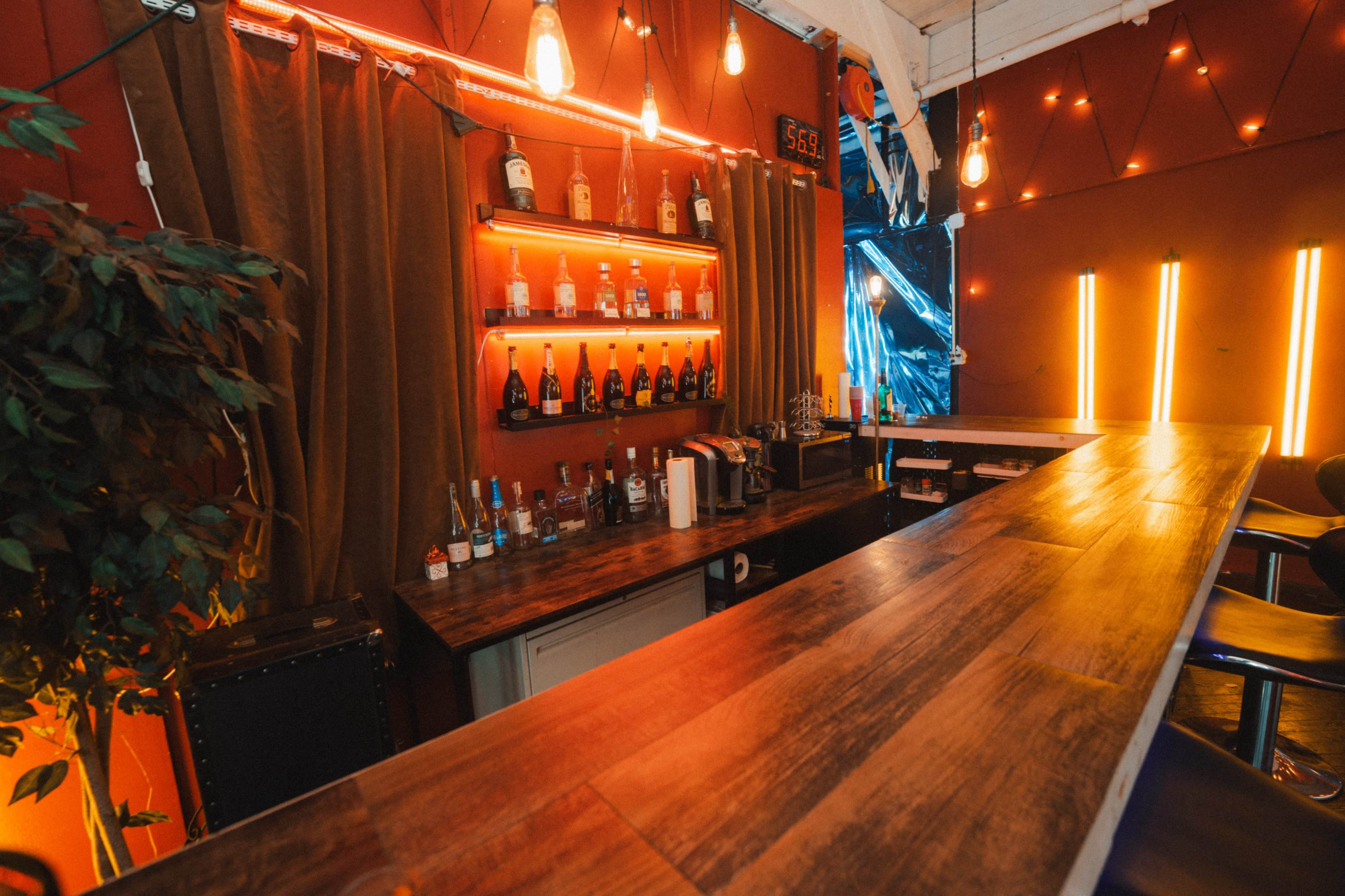 The image shows a well-lit bar area with a wooden counter, shelves displaying various bottles, and warm lighting accents.