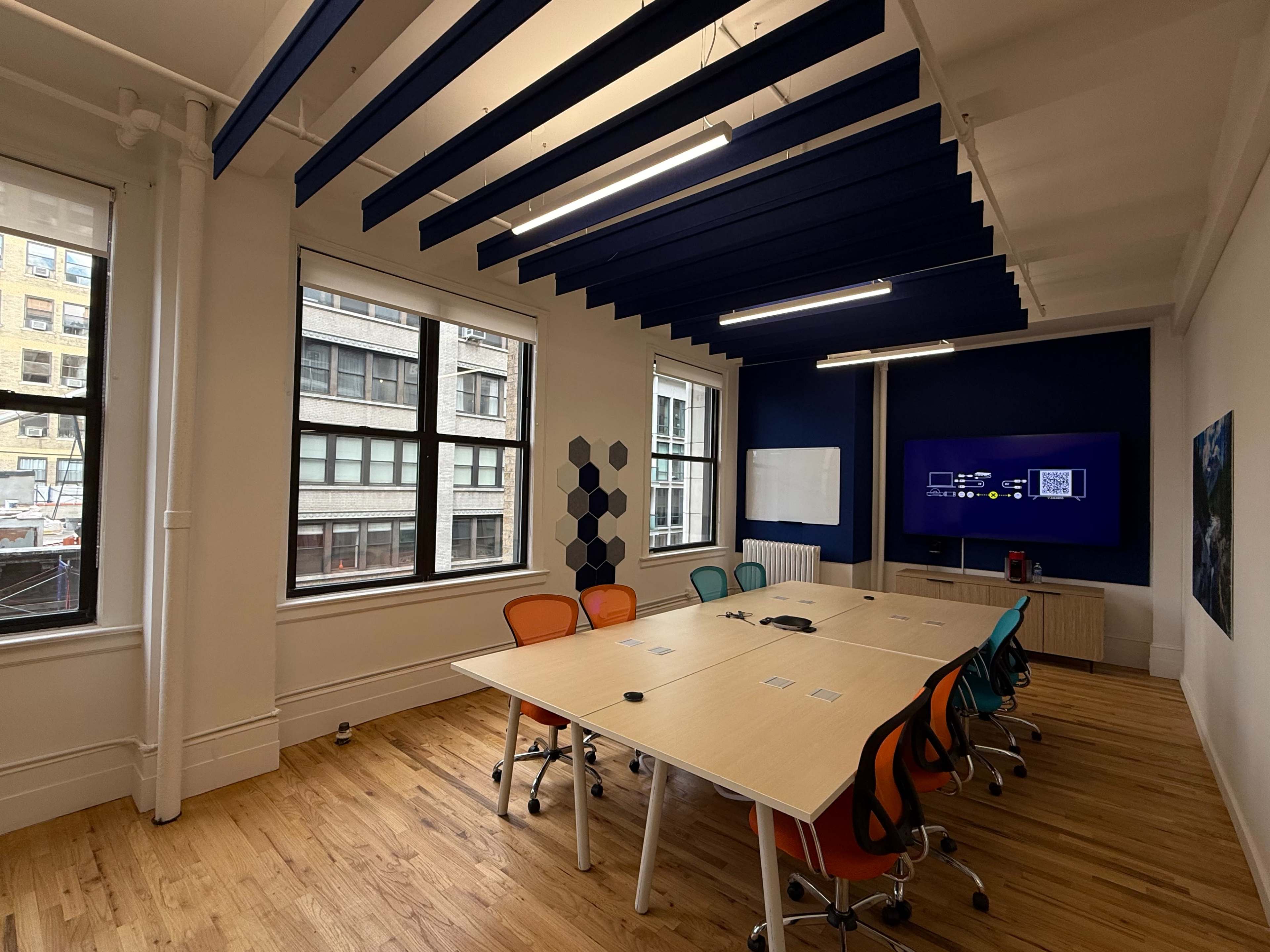 A modern conference room features a large table surrounded by colorful chairs, with windows providing natural light and a display screen mounted on the wall.
