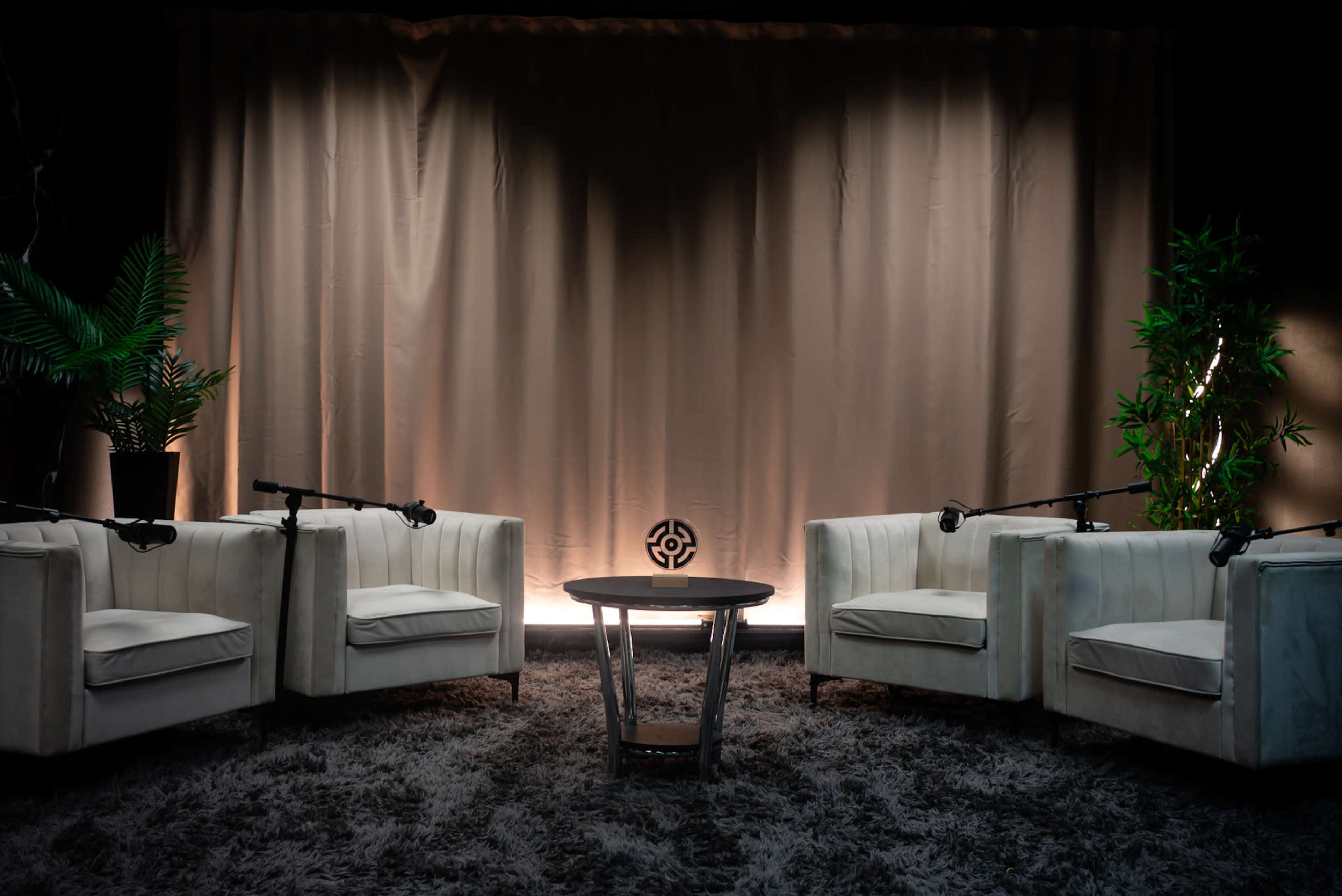 The image shows a minimalist interview setup with three white armchairs, a round table, and lush green plants in a dimly lit space.