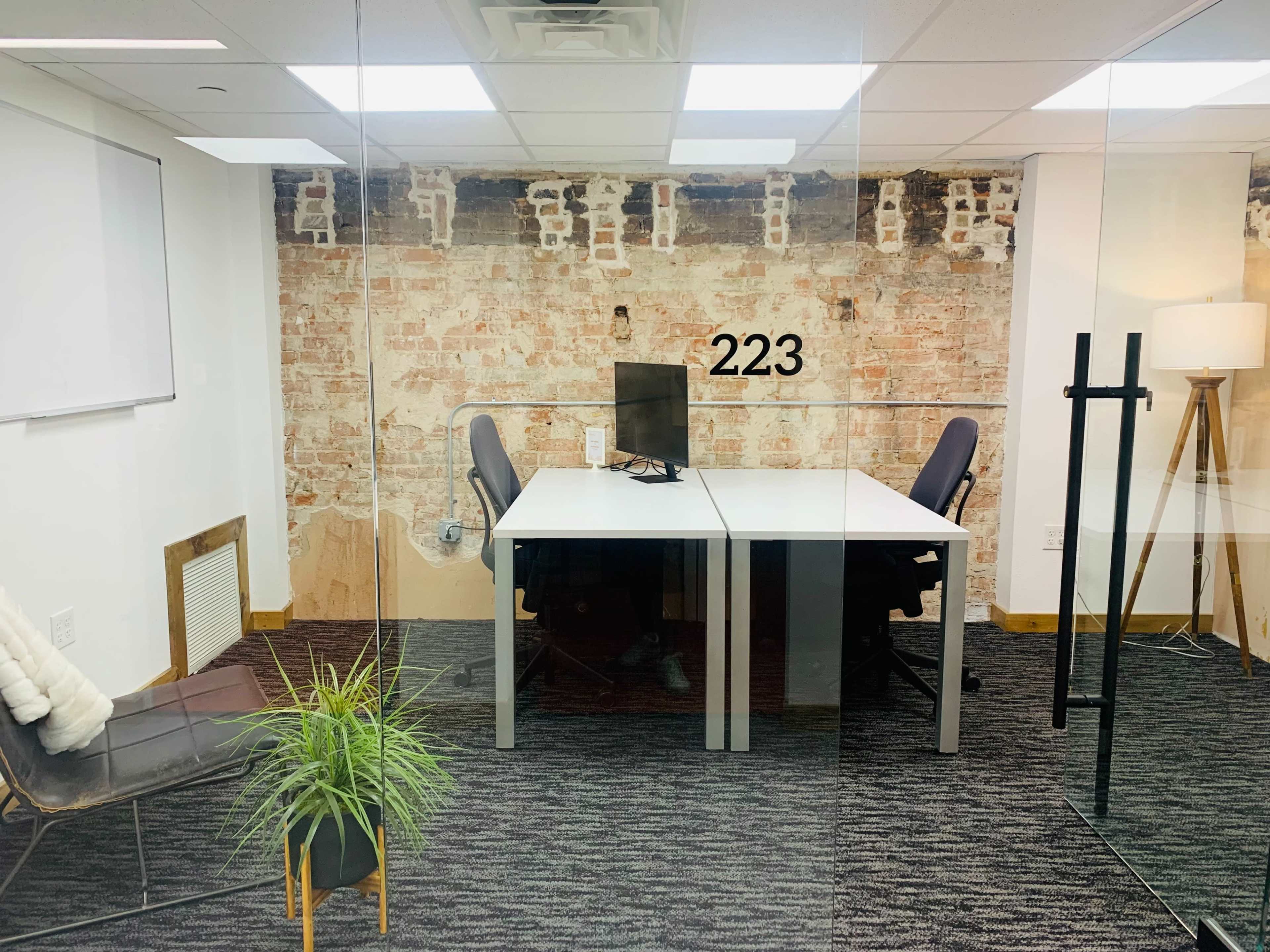 A glass-walled office features two desk chairs, a computer monitor, and exposed brick walls with the number 223 displayed.