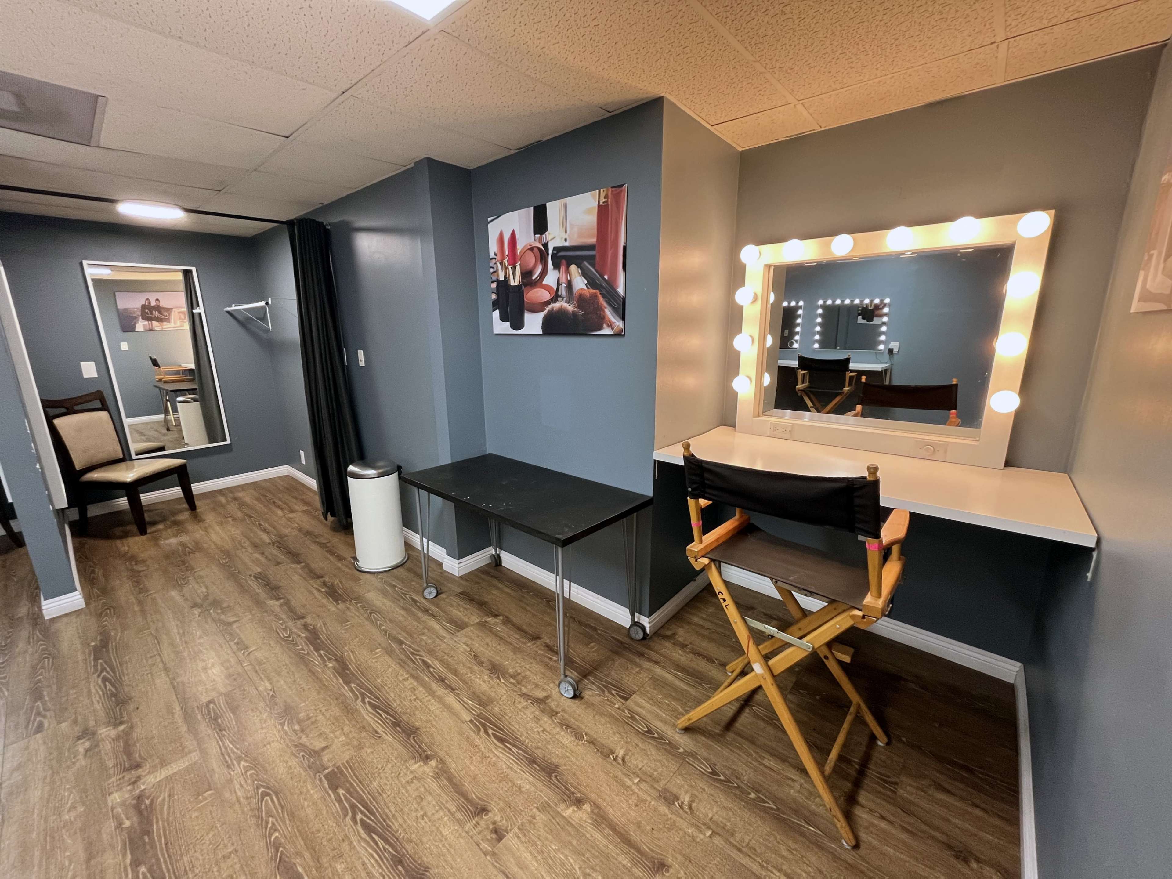 A well-lit dressing room featuring a mirror with lights, a makeup chair, a folding table, and a mirror on the back wall.