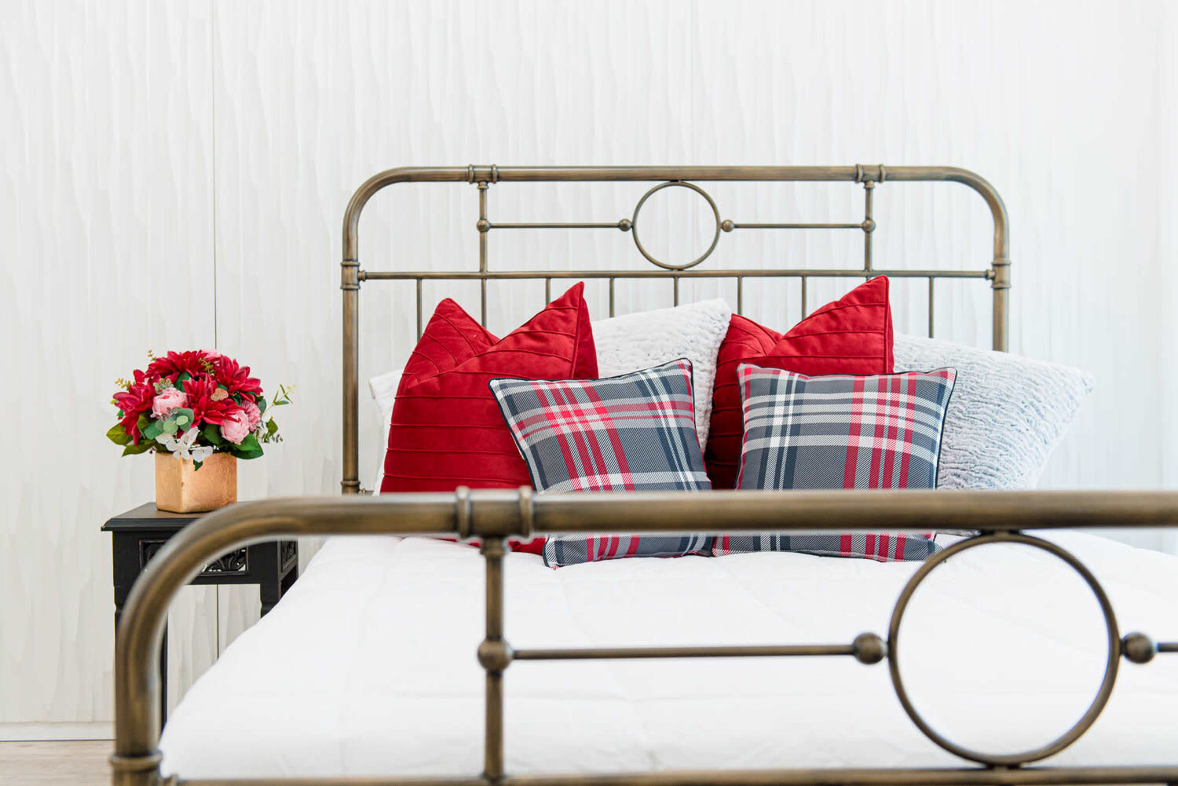 The image features a neatly made bed with a vintage metal frame, adorned with red and plaid pillows, and a small vase of flowers on a nightstand nearby.