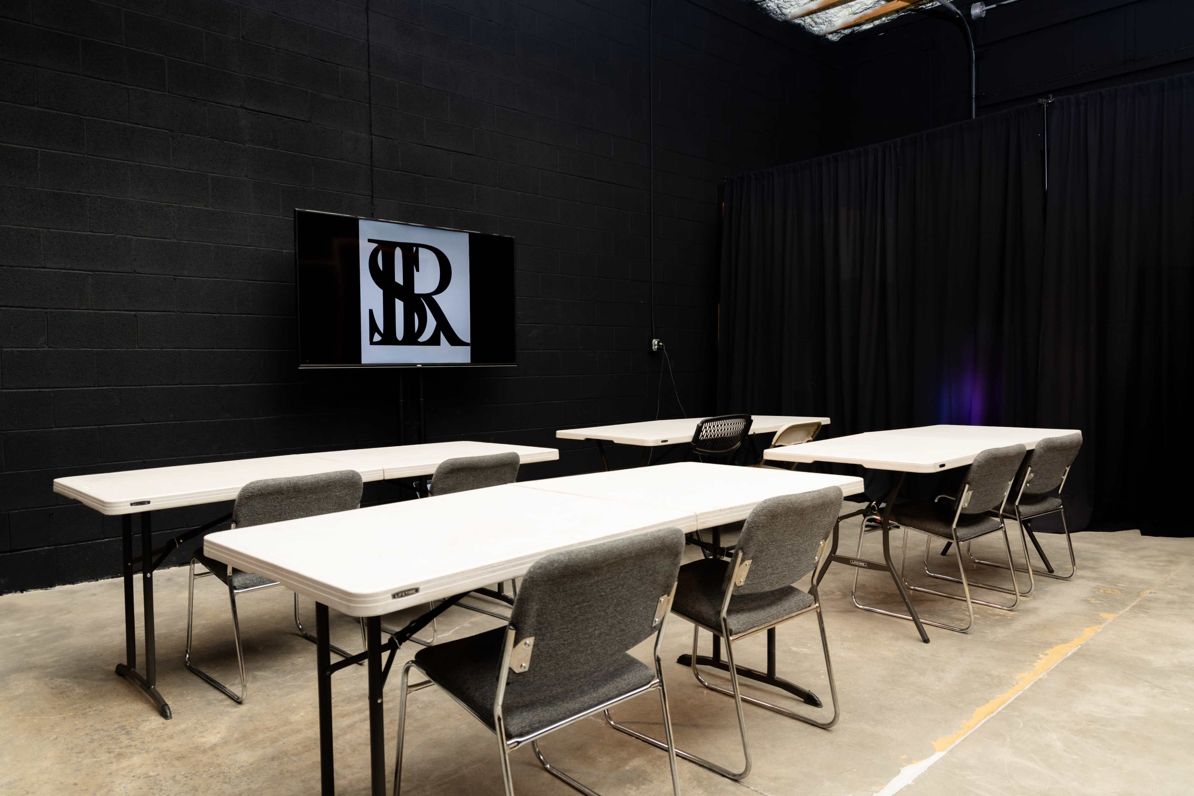The image shows a seminar room with several rectangular tables and chairs arranged facing a screen displaying a logo, against a backdrop of black walls and curtains.