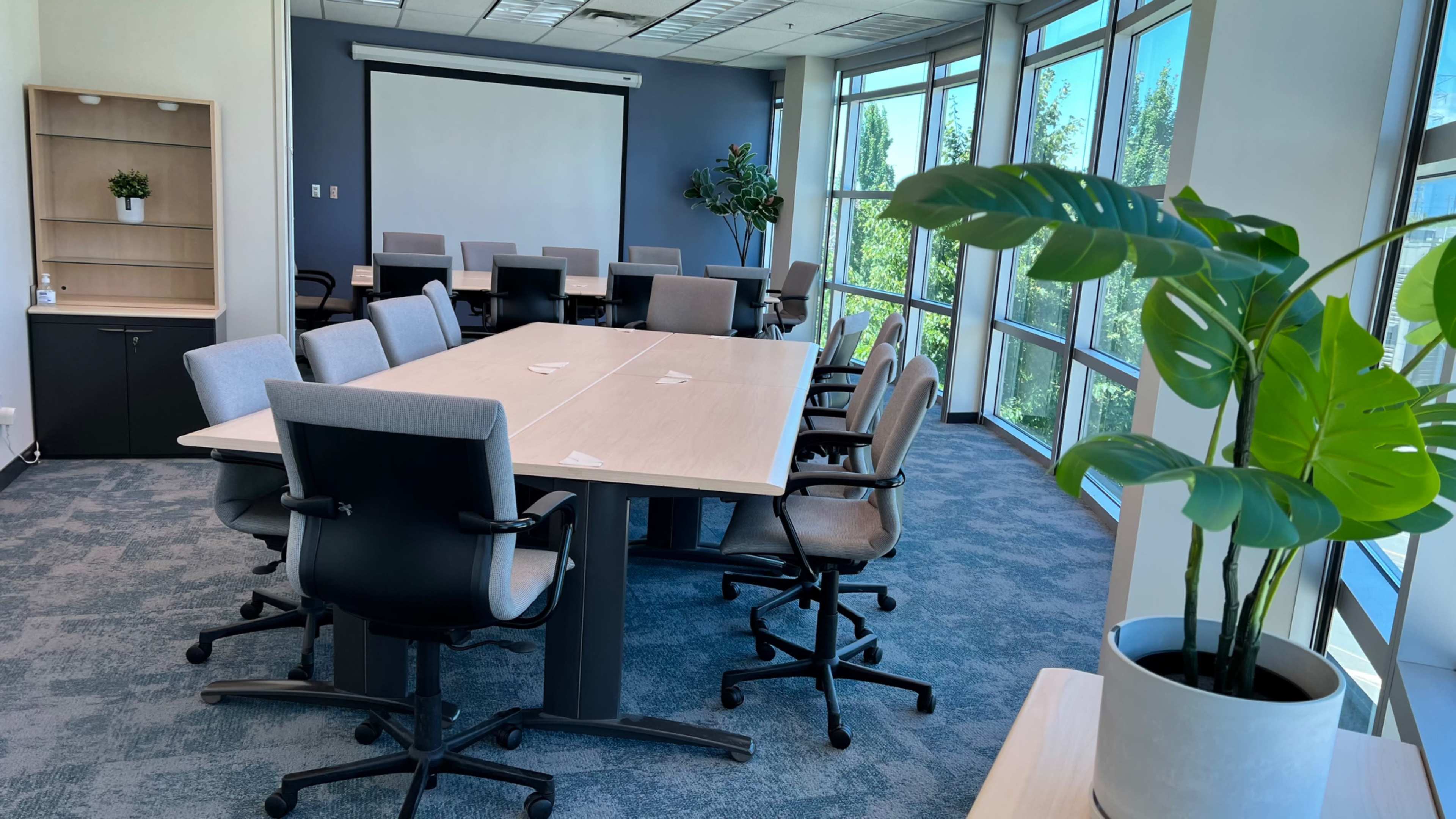 A spacious conference room features a large table surrounded by ergonomic chairs, with a projector screen and windows that provide ample natural light.
