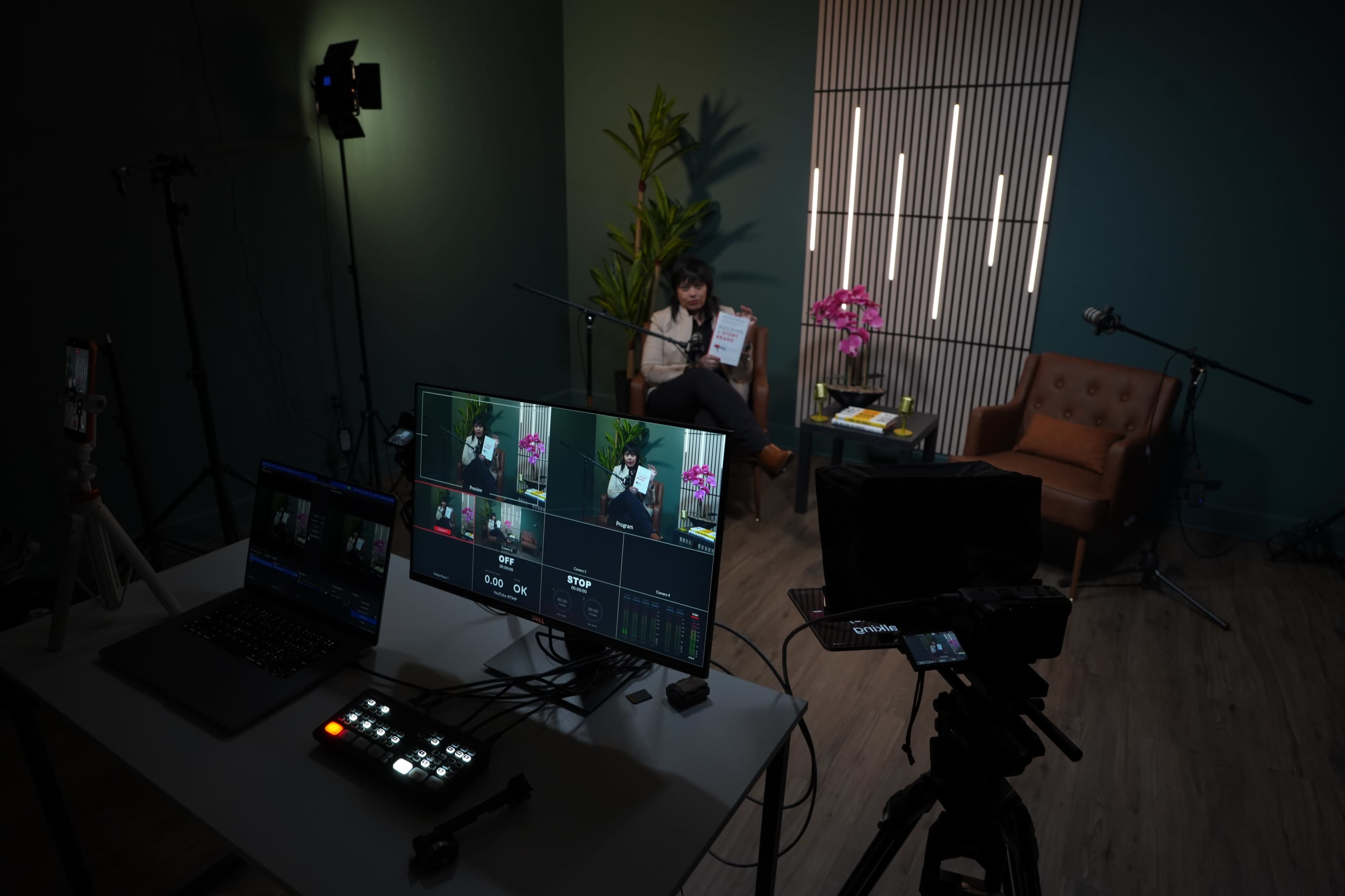 A woman sits in a chair holding a document in a well-lit studio with cameras and lighting equipment set up around her.