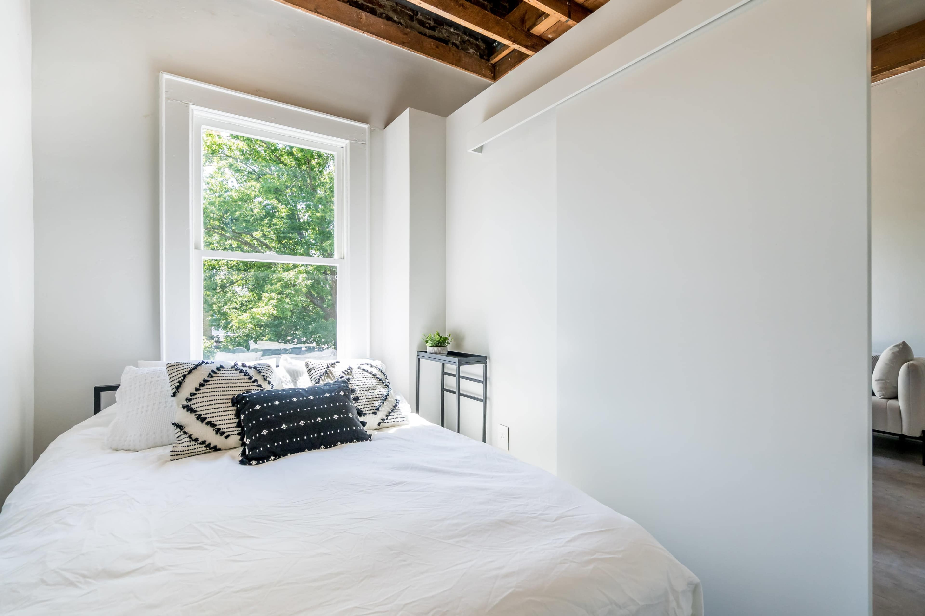 A minimalist bedroom with a bed covered in white linens and decorative pillows, next to a large window that allows natural light to enter.