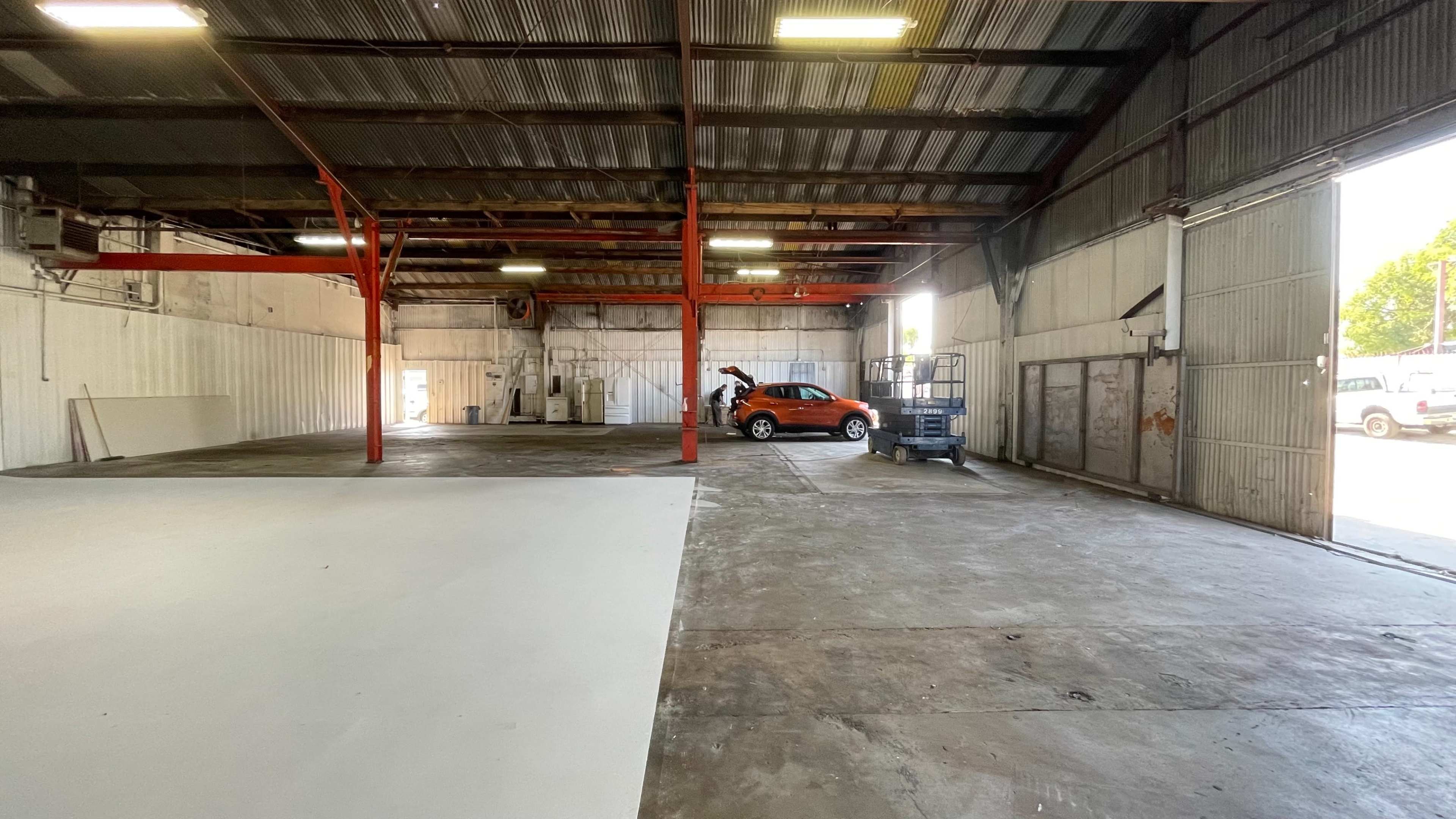 An industrial warehouse interior features a parked orange vehicle, a scissor lift, and open space with exposed beams and concrete flooring.