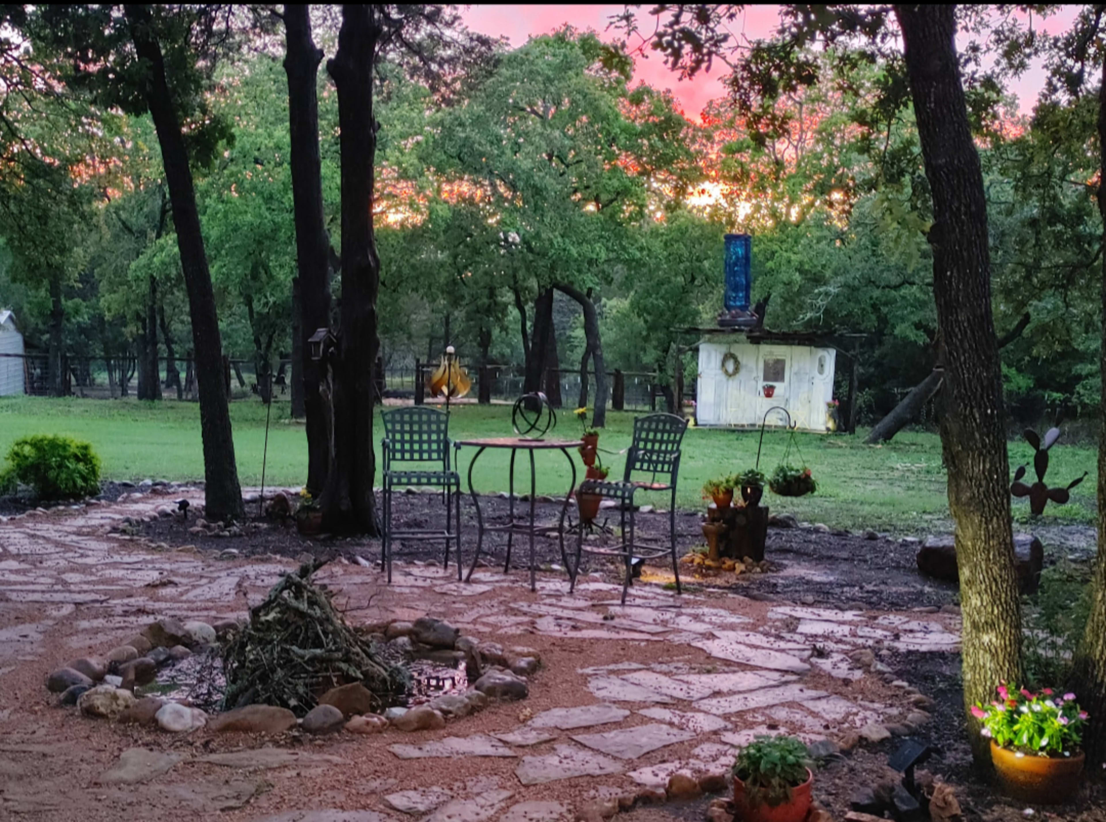 A circular stone fire pit is surrounded by a patio area with a table and chairs, set against a backdrop of trees and a rustic shed under a colorful sunset sky.