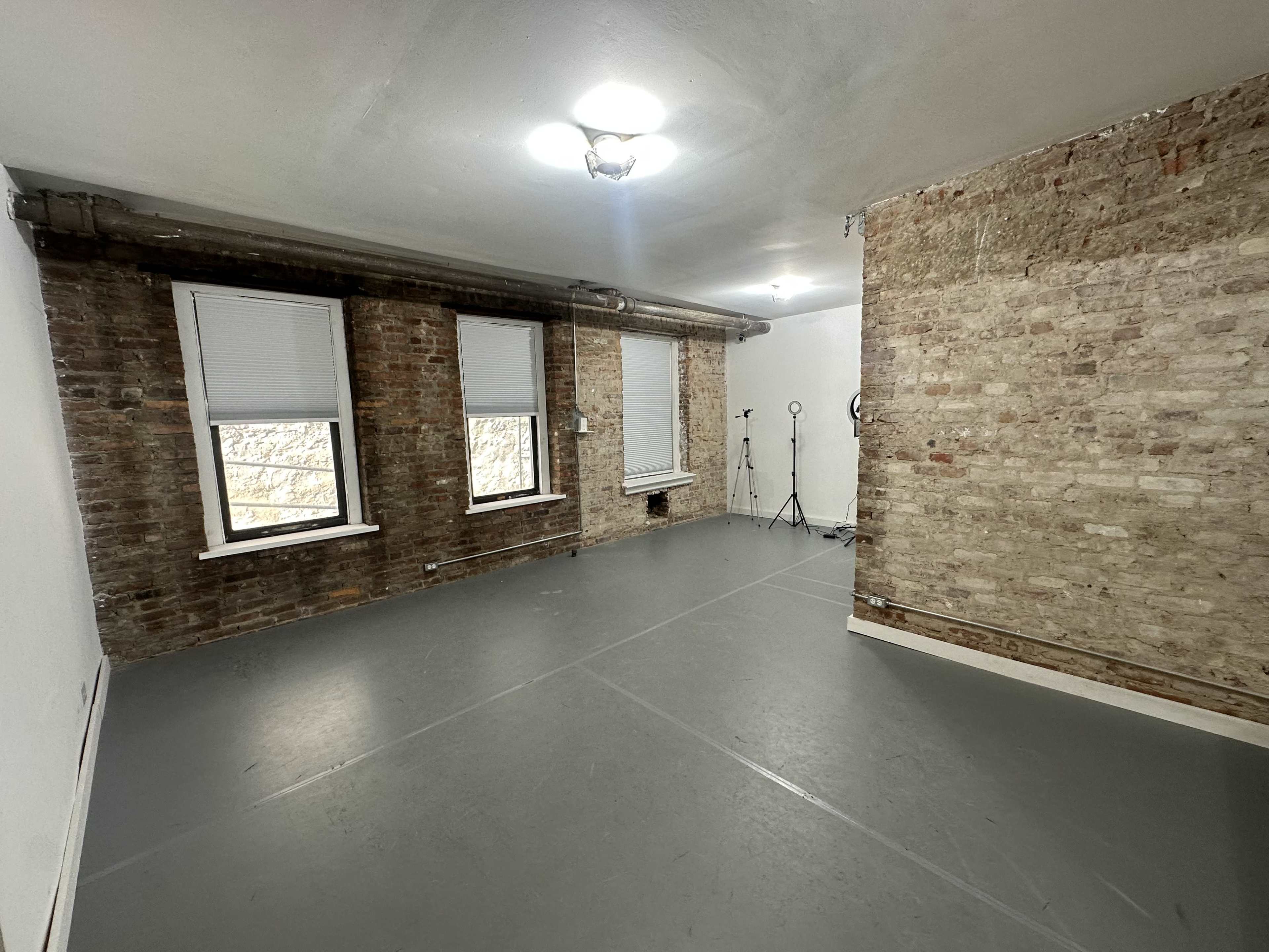 A spacious room with exposed brick walls, large windows with blinds, and a gray floor, featuring a minimalistic setup in one corner.