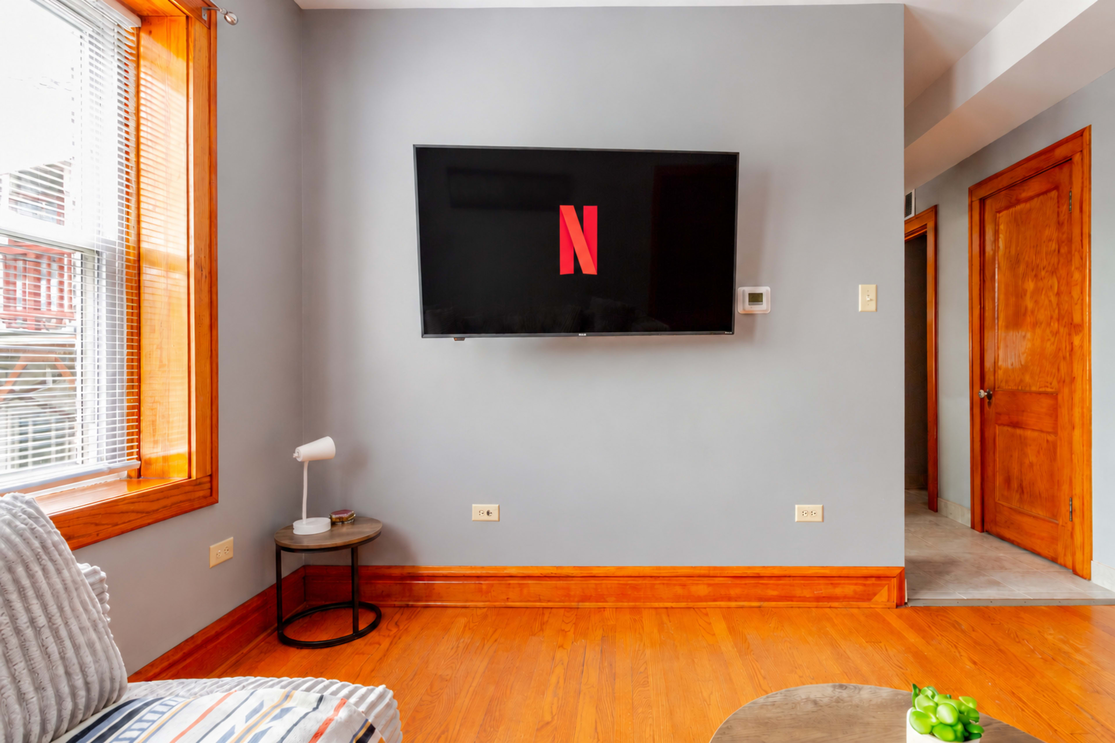 A flat-screen television displaying the Netflix logo is mounted on a gray wall in a room with wooden accents and a doorway visible to the right.