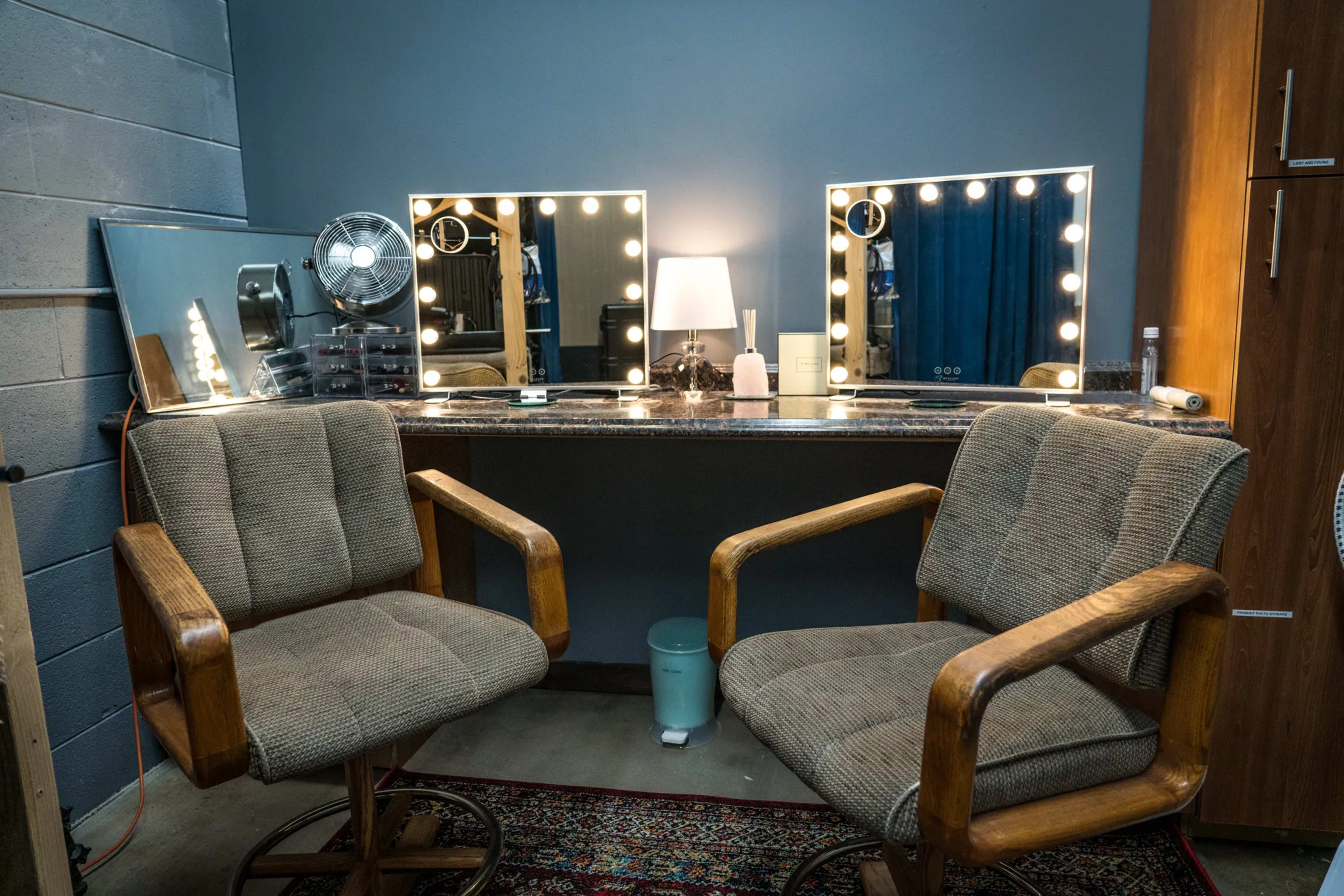 A salon-style vanity area with two wooden chairs facing a mirrored countertop illuminated by bright vanity lights, alongside a fan and various grooming products.
