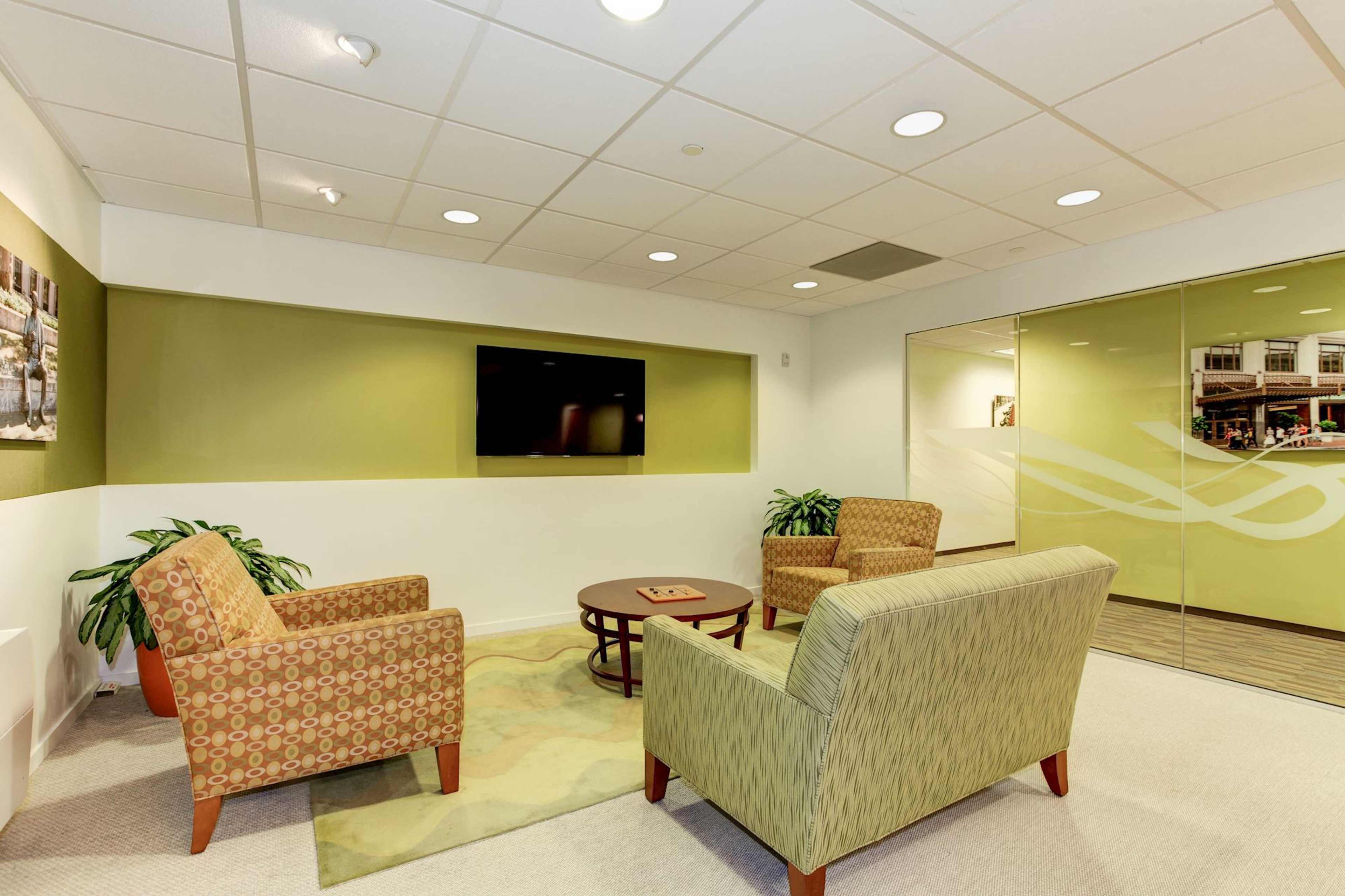A small lounge area with two patterned armchairs, a round table, and a wall-mounted television, surrounded by green accents and a carpet.