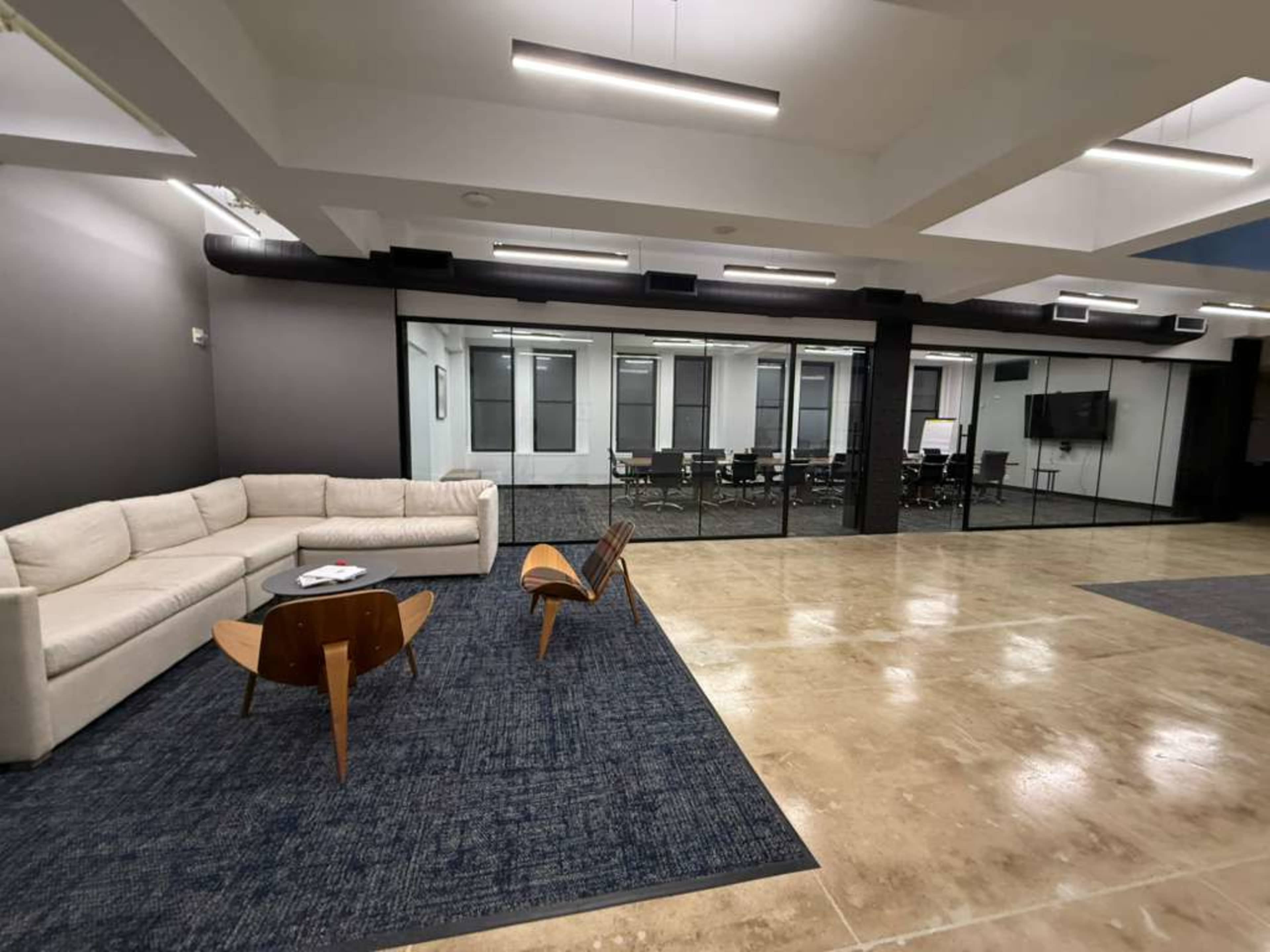 The image shows a modern office lounge area with a sofa, a coffee table, and a view of a glass-walled conference room.