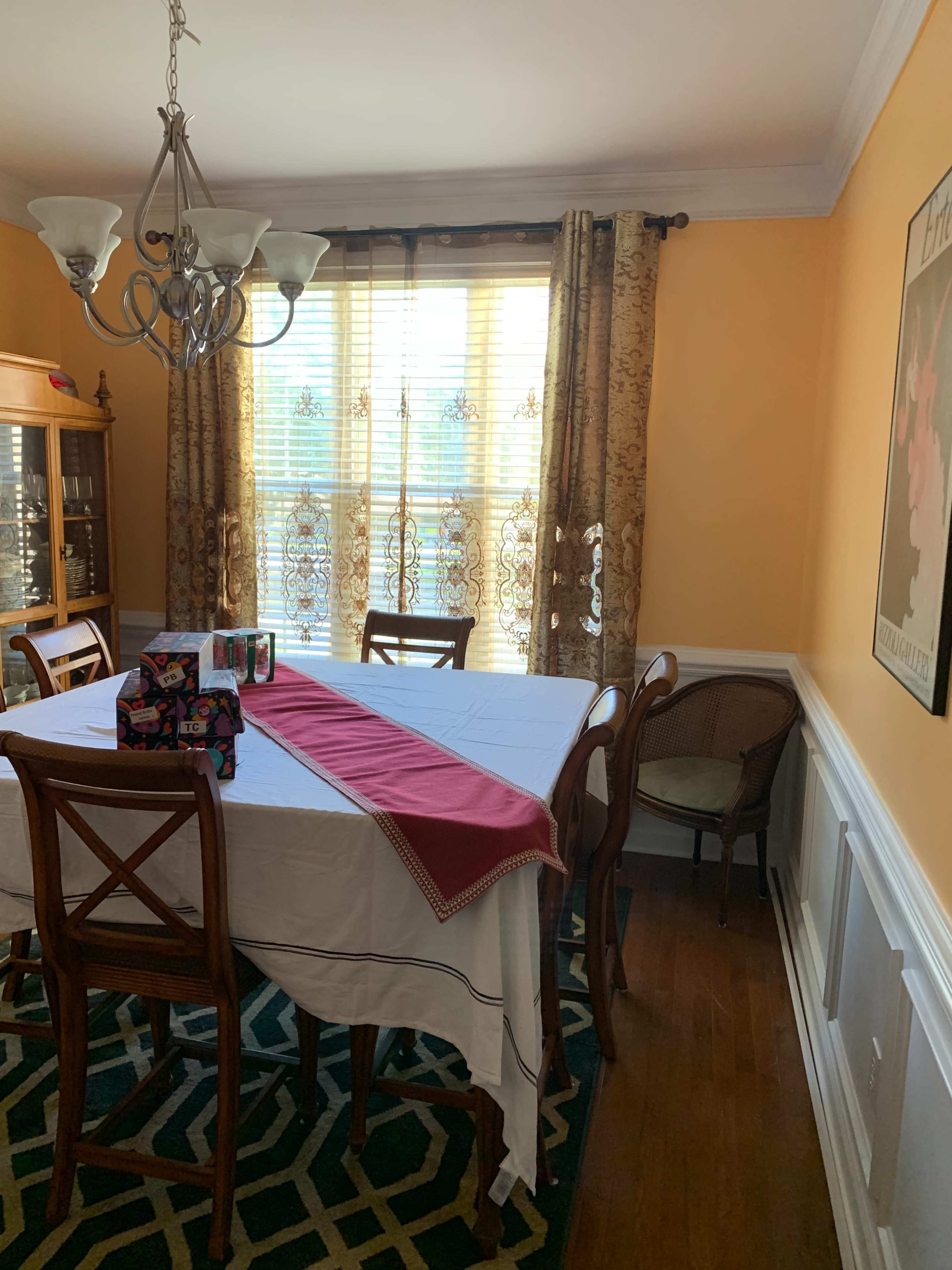 A dining room with a table covered by a white tablecloth, a red runner down the center, and several wrapped gifts on the table, surrounded by wooden chairs and a window with patterned curtains.