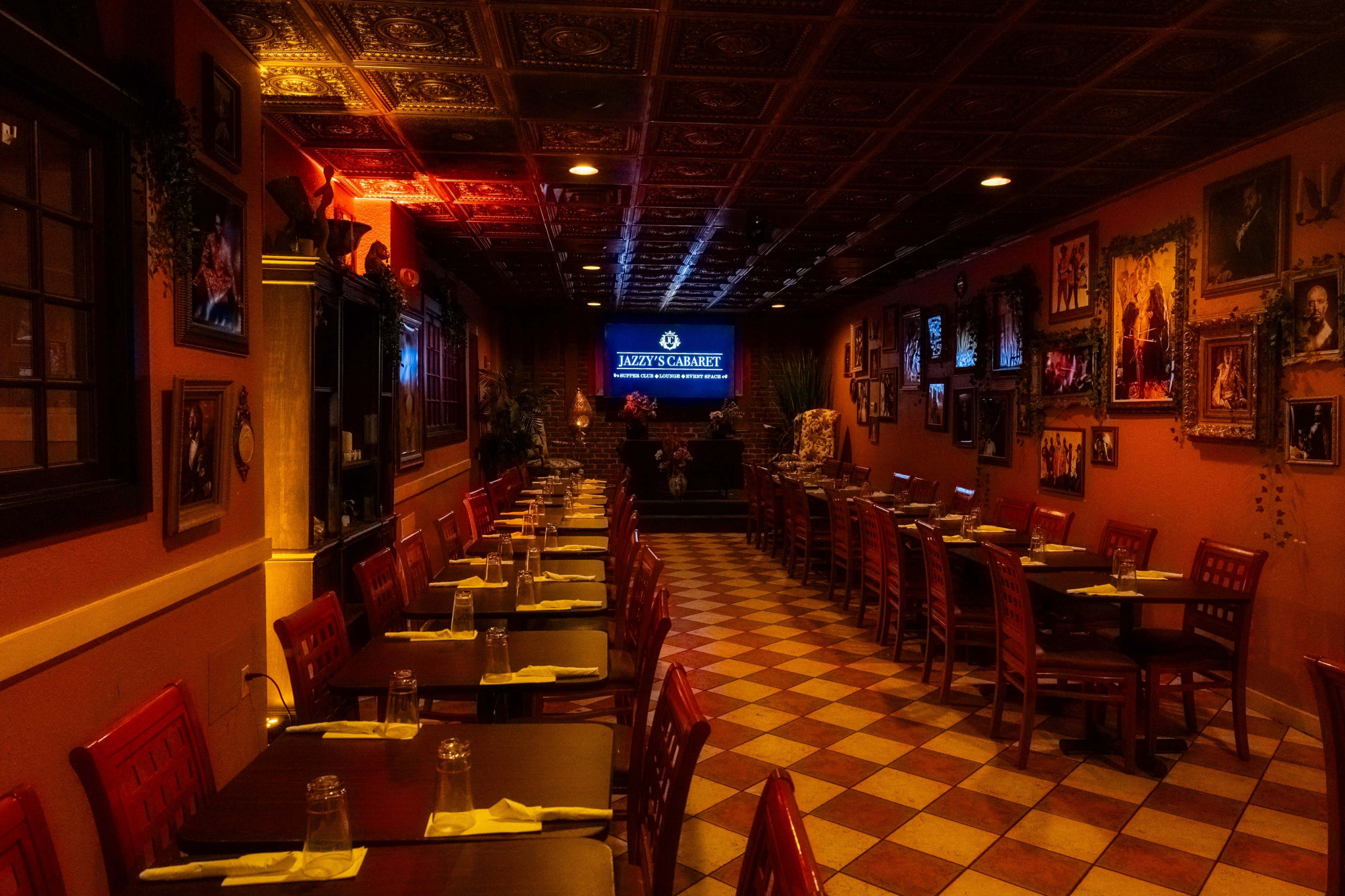 An empty restaurant interior featuring red wooden chairs and tables, a decorative ceiling, and walls adorned with framed photographs.