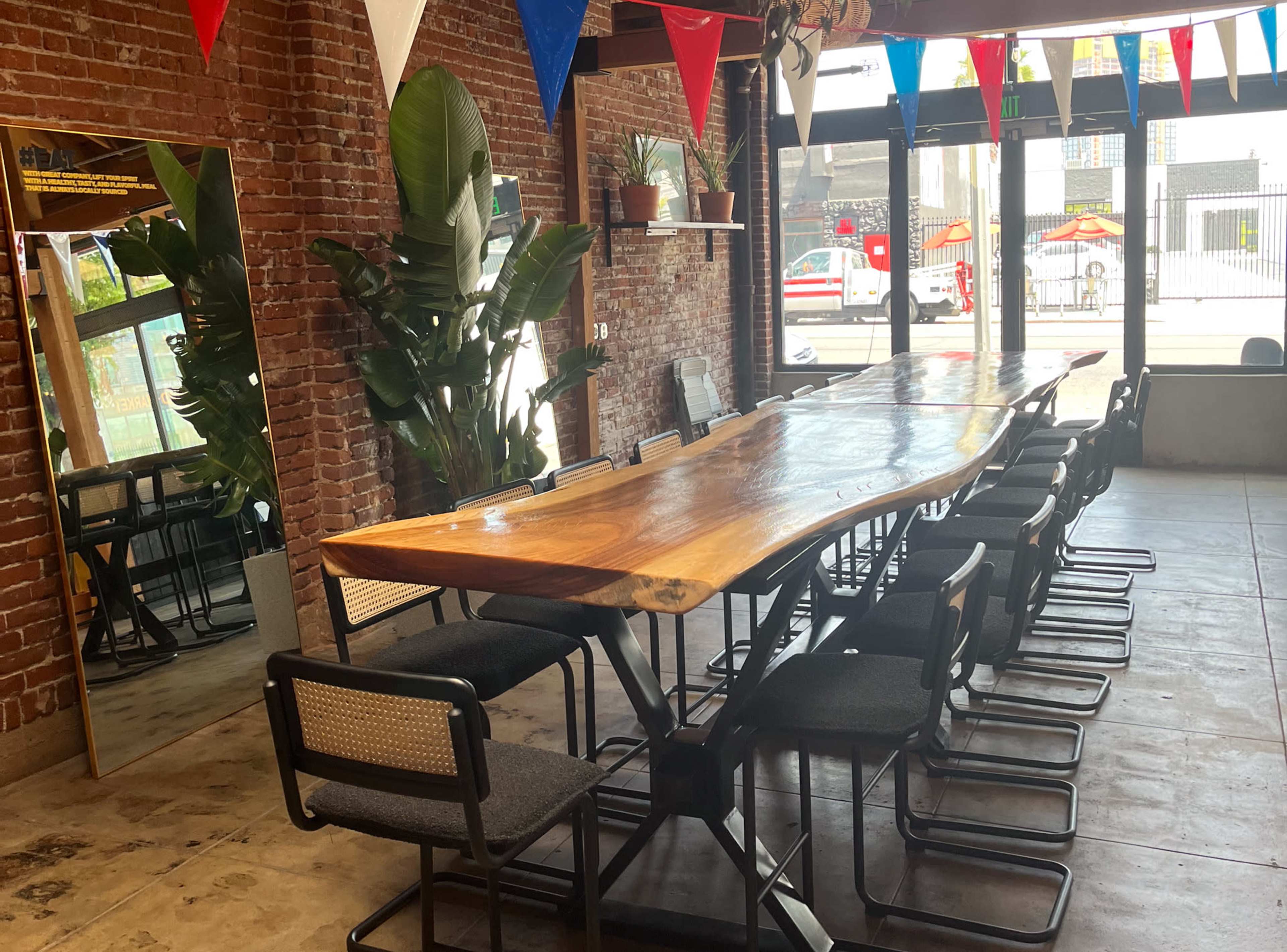 A long wooden table with chairs is set against a backdrop of exposed brick walls and large windows, decorated with colorful triangular flags.