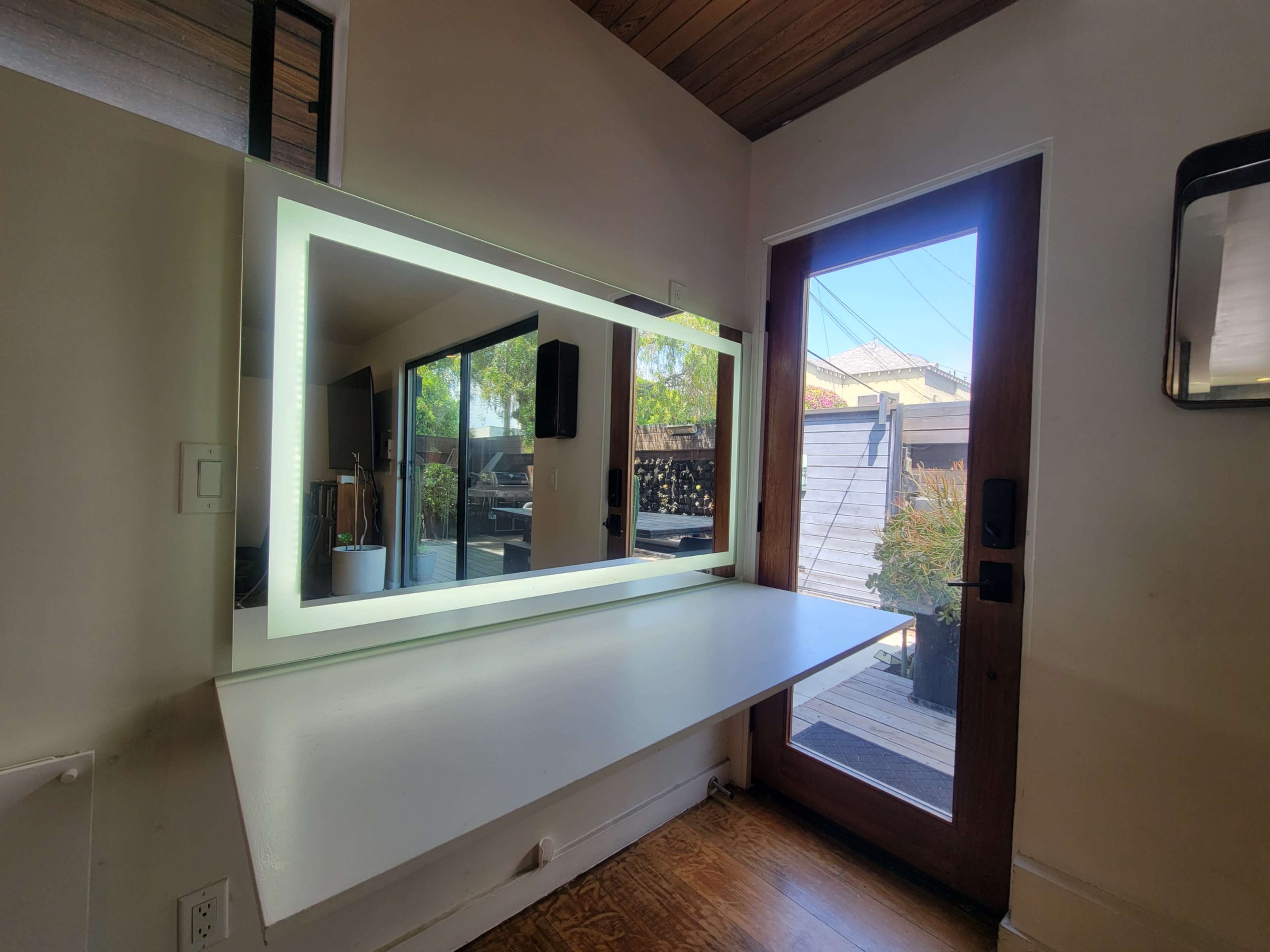 A modern workspace features a large illuminated mirror mounted on the wall, with a door leading to an outdoor area visible beside it.