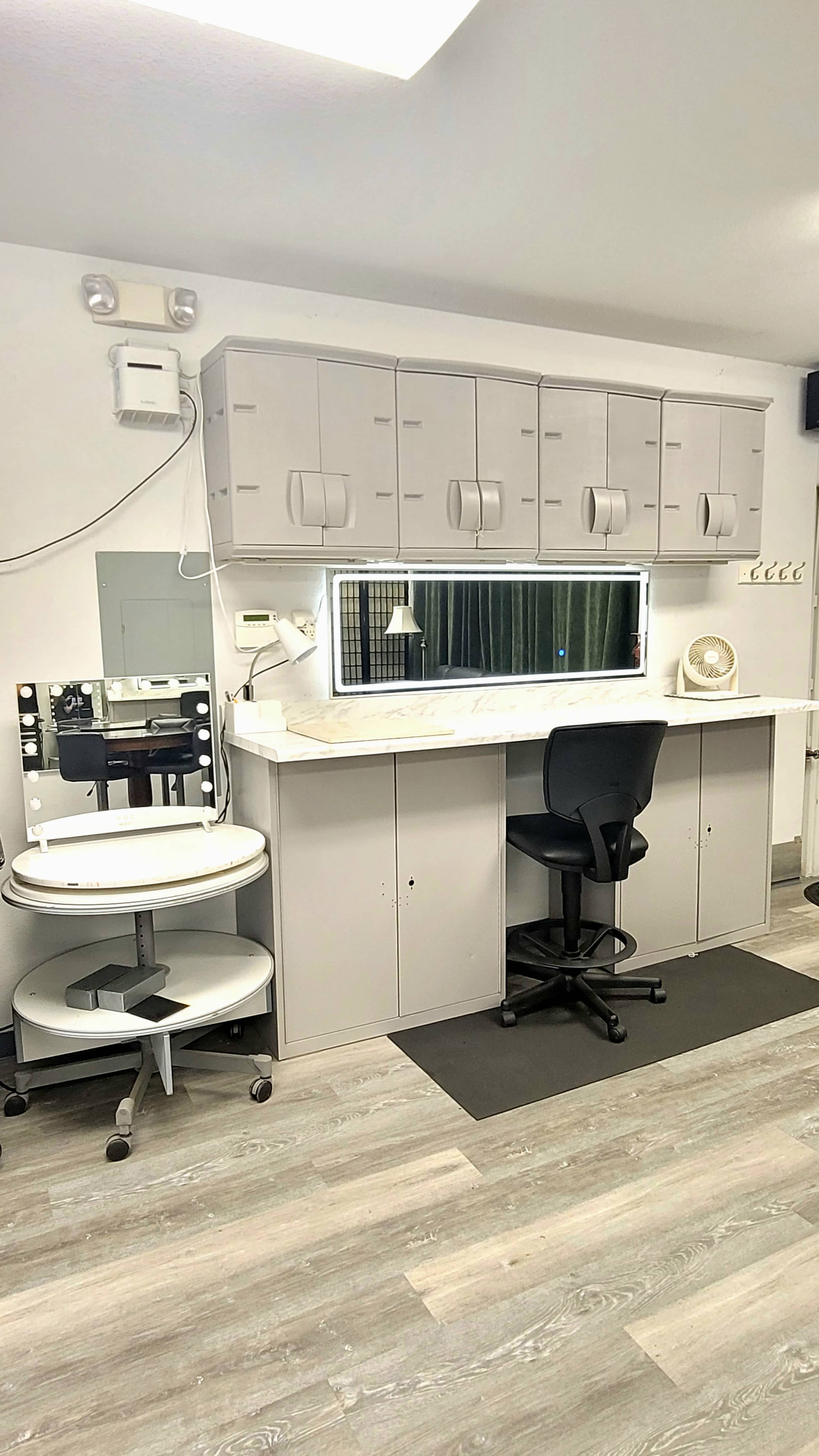 A minimalist beauty or grooming station with a large mirror, a rolling cart, and a black chair against a light-colored wall.