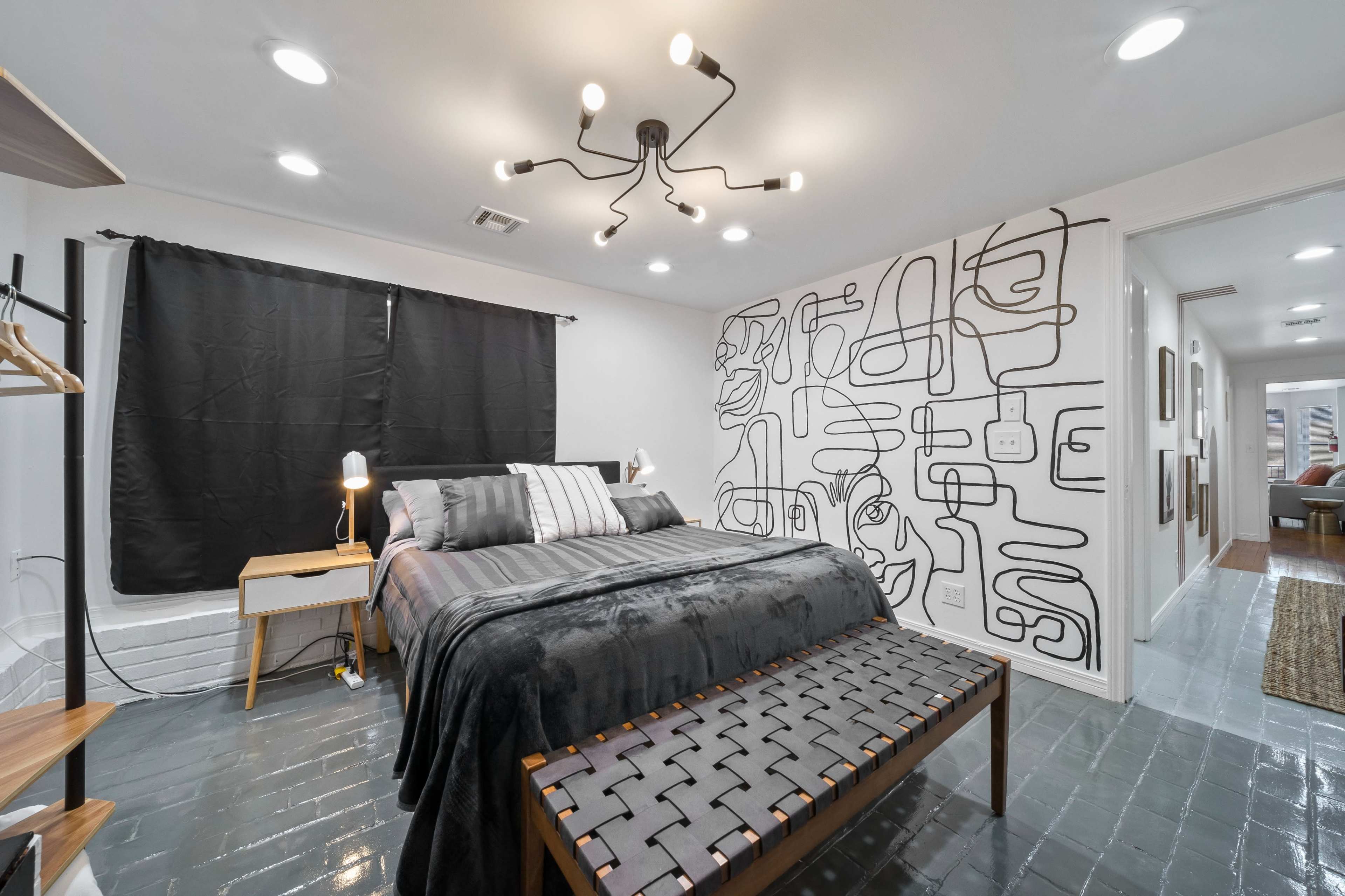 A modern bedroom features a bed with gray bedding, a bench at the foot, and a large abstract mural on one wall.