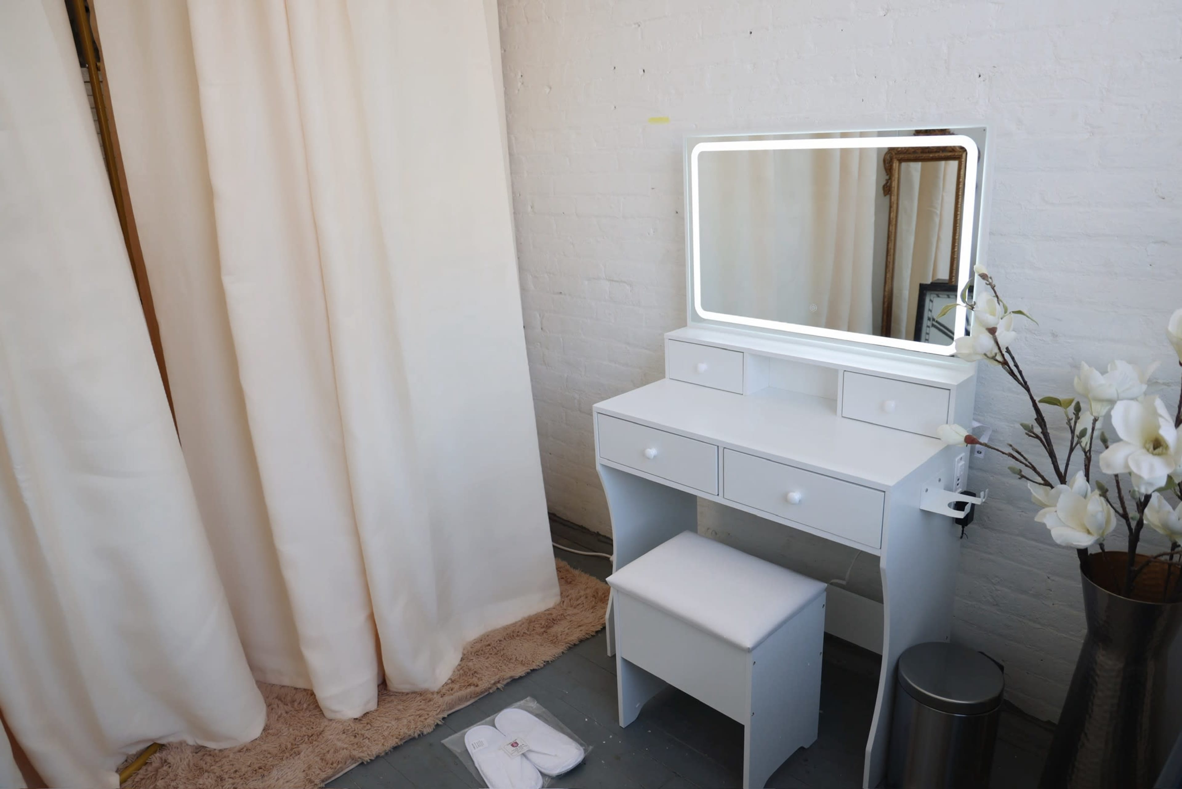 The image features a white vanity table with a mirror, two drawers, and a stool, situated next to a curtain and a potted plant.