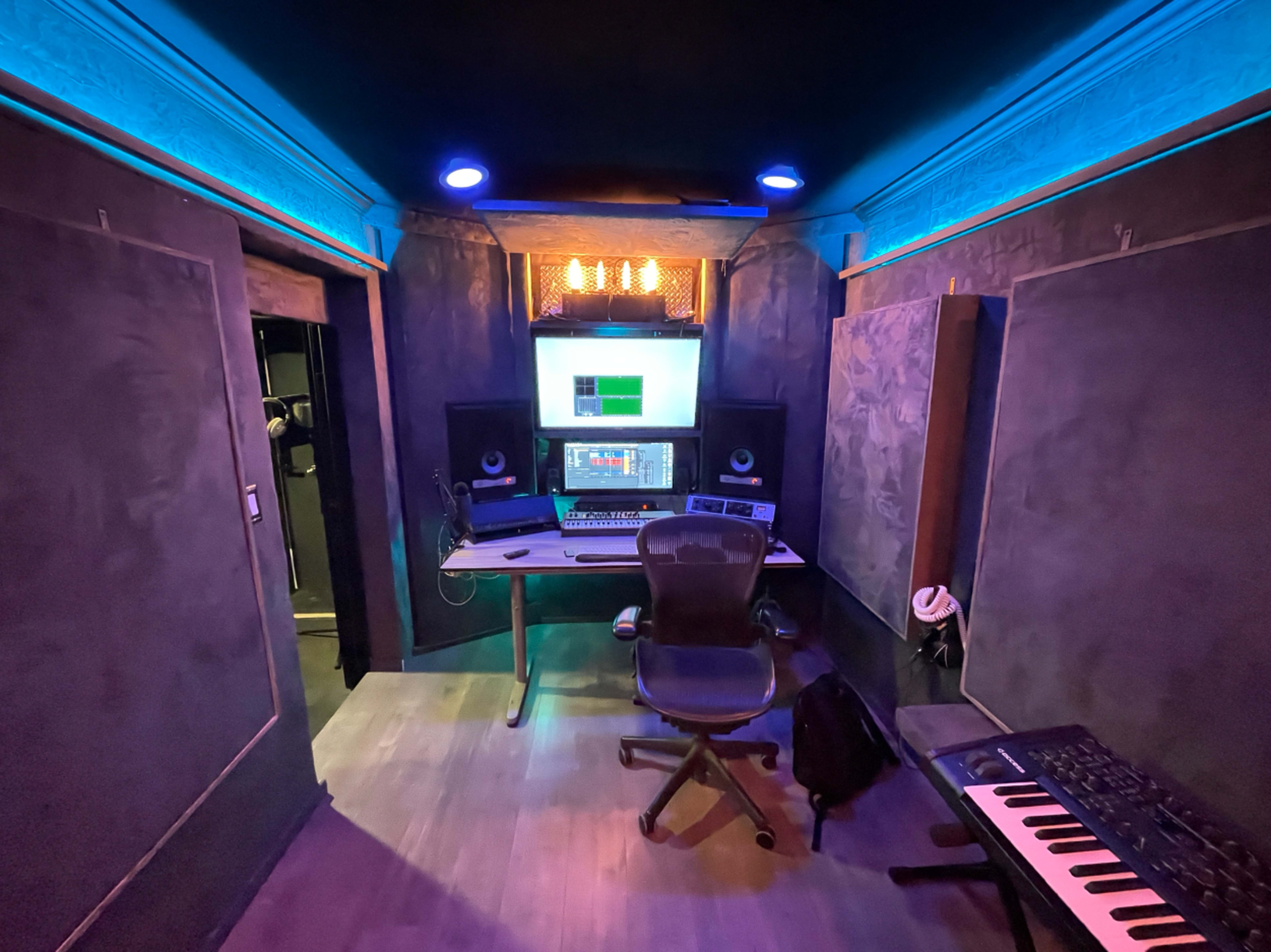 A music recording studio features a central workstation with a computer, audio equipment, and soundproof walls illuminated by blue and amber lights.