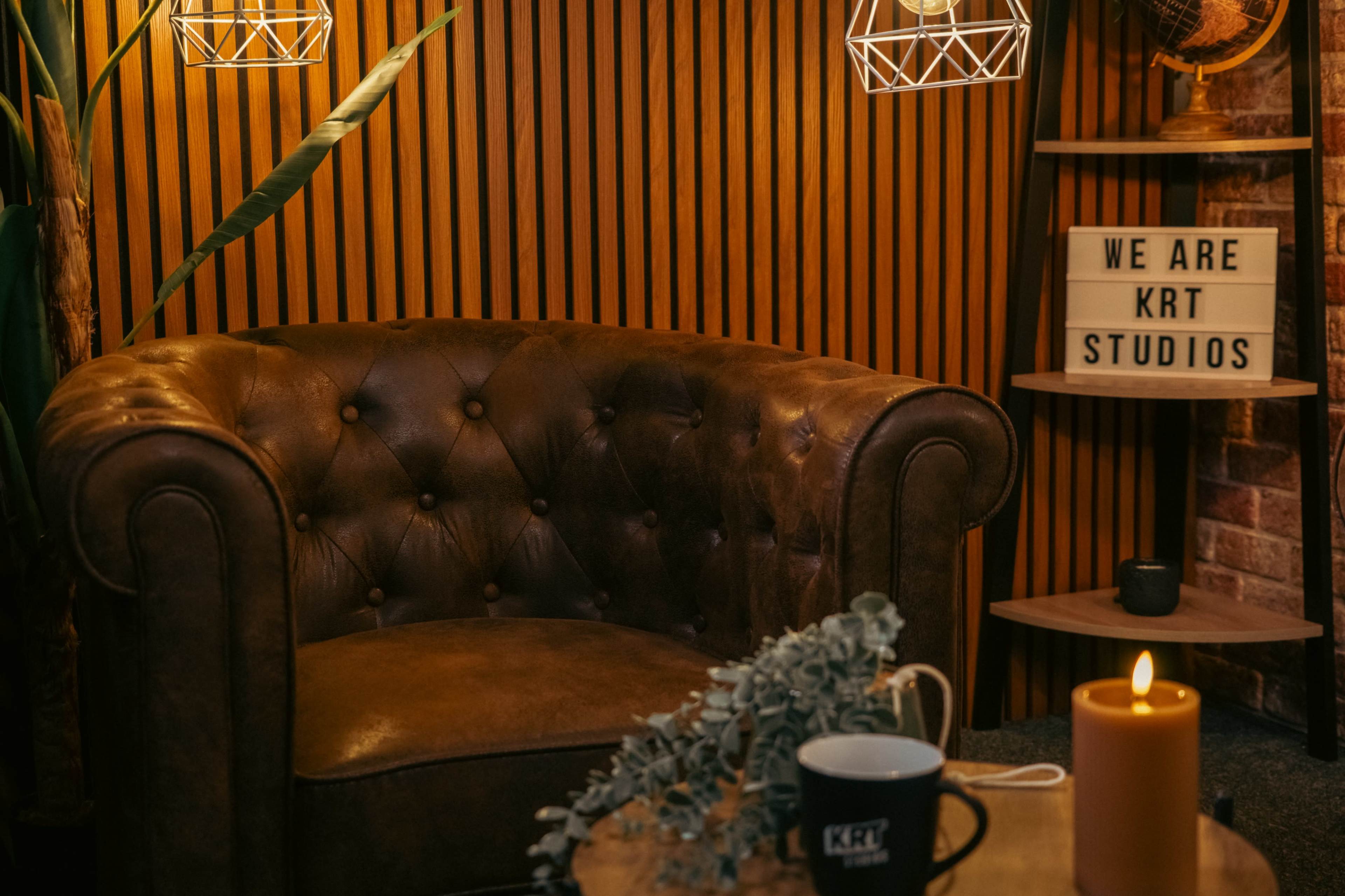 The image features a cozy corner with a tufted leather armchair, a wooden table with a plant, and a lit candle, alongside decorative signage that reads "WE ARE KRT STUDIOS."