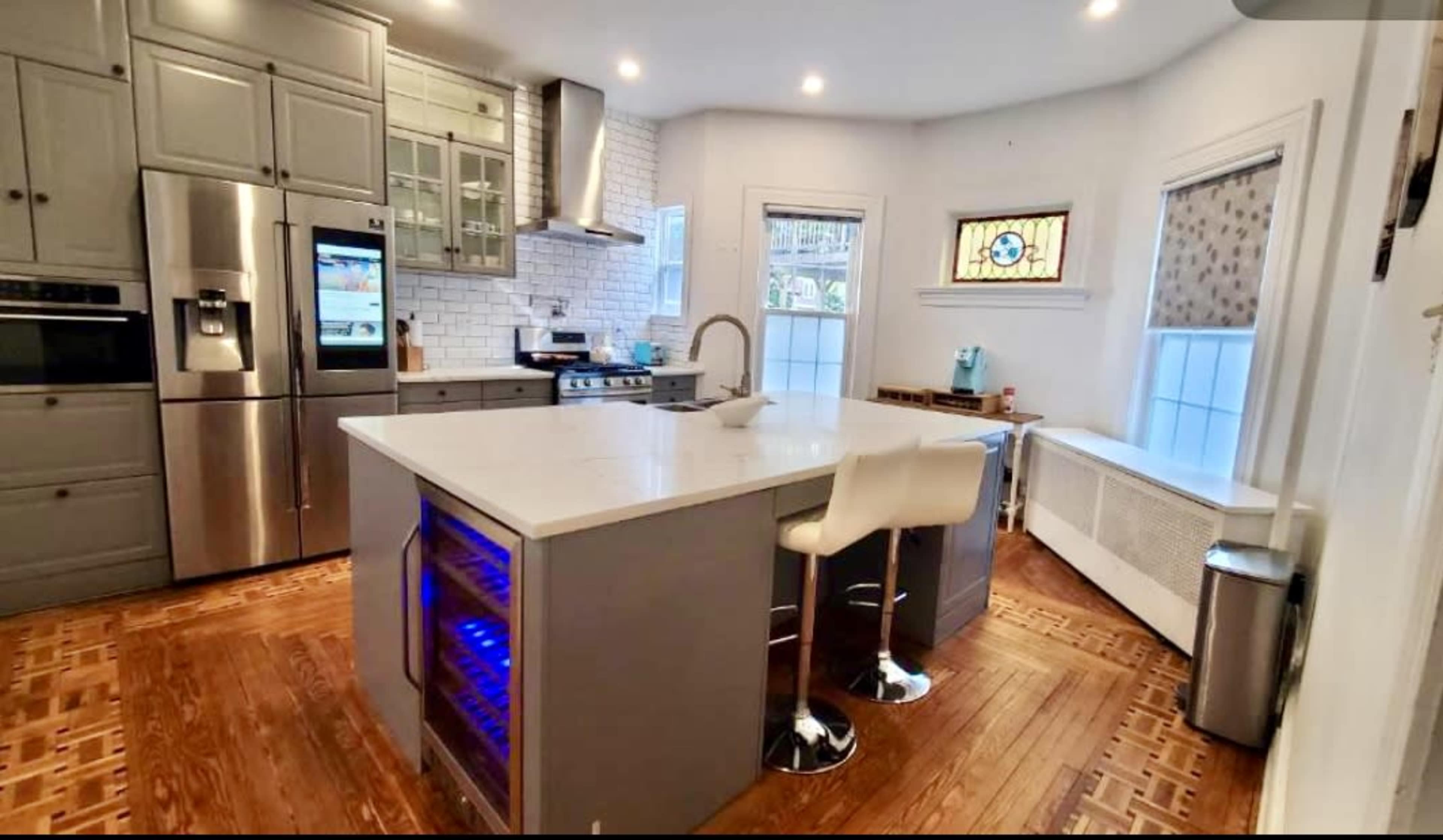 The image shows a modern kitchen featuring gray cabinetry, stainless steel appliances, a large island with seating, and hardwood flooring.