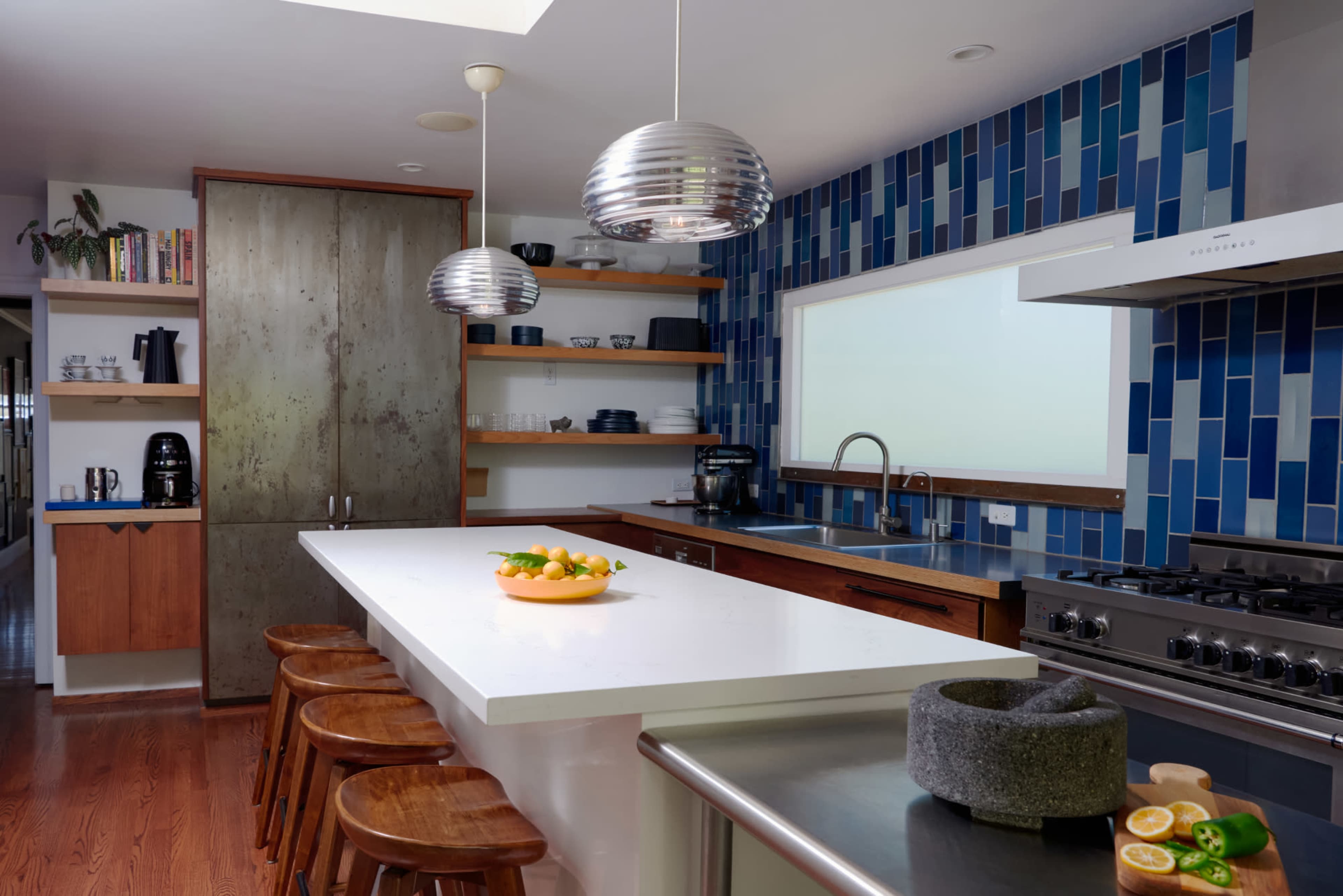 A modern kitchen features a white island countertop, wooden bar stools, blue tiled walls, and stainless steel appliances.