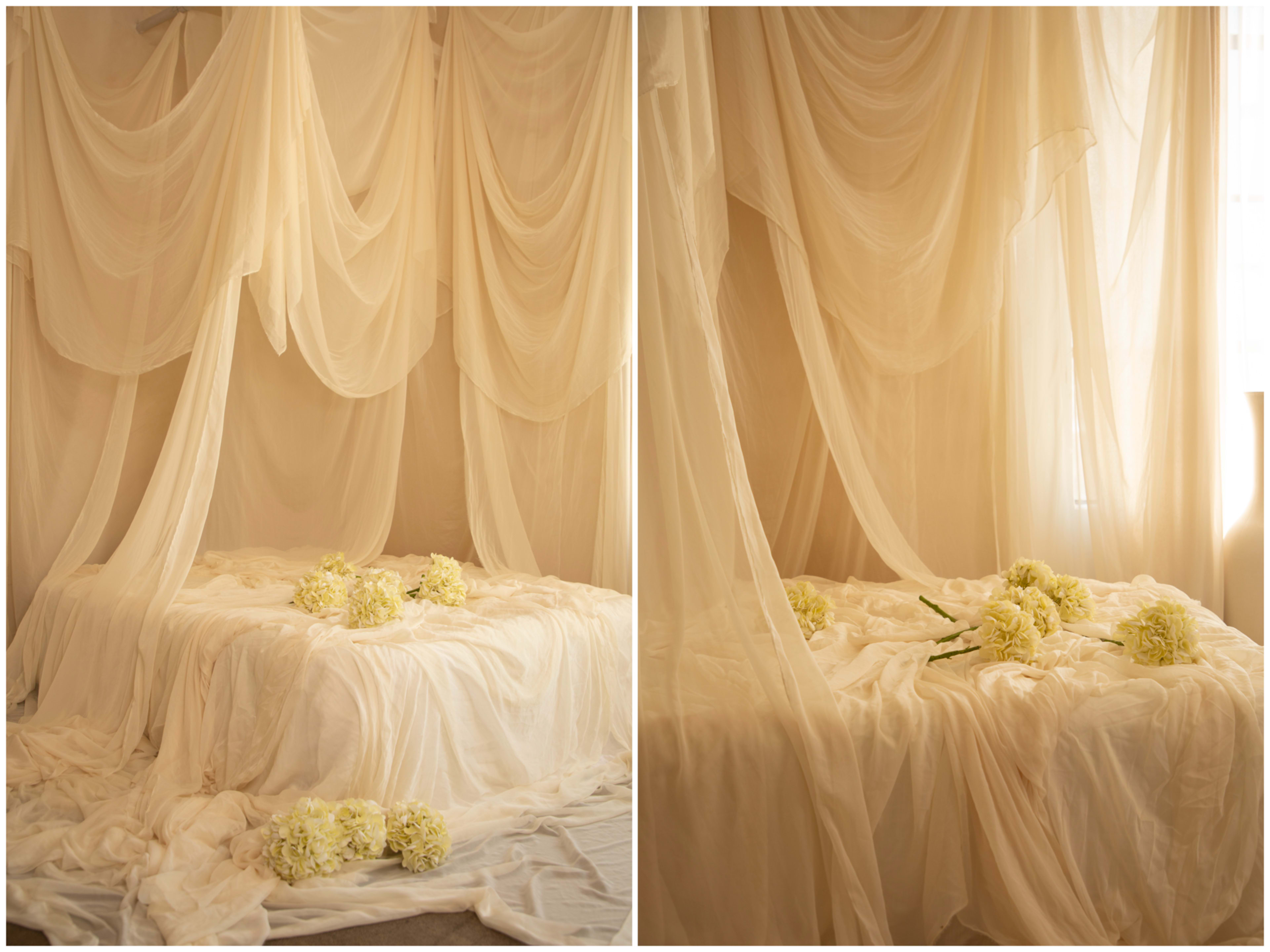 The image shows a softly lit bedroom scene with a bed draped in layers of sheer fabric and adorned with white flowers.