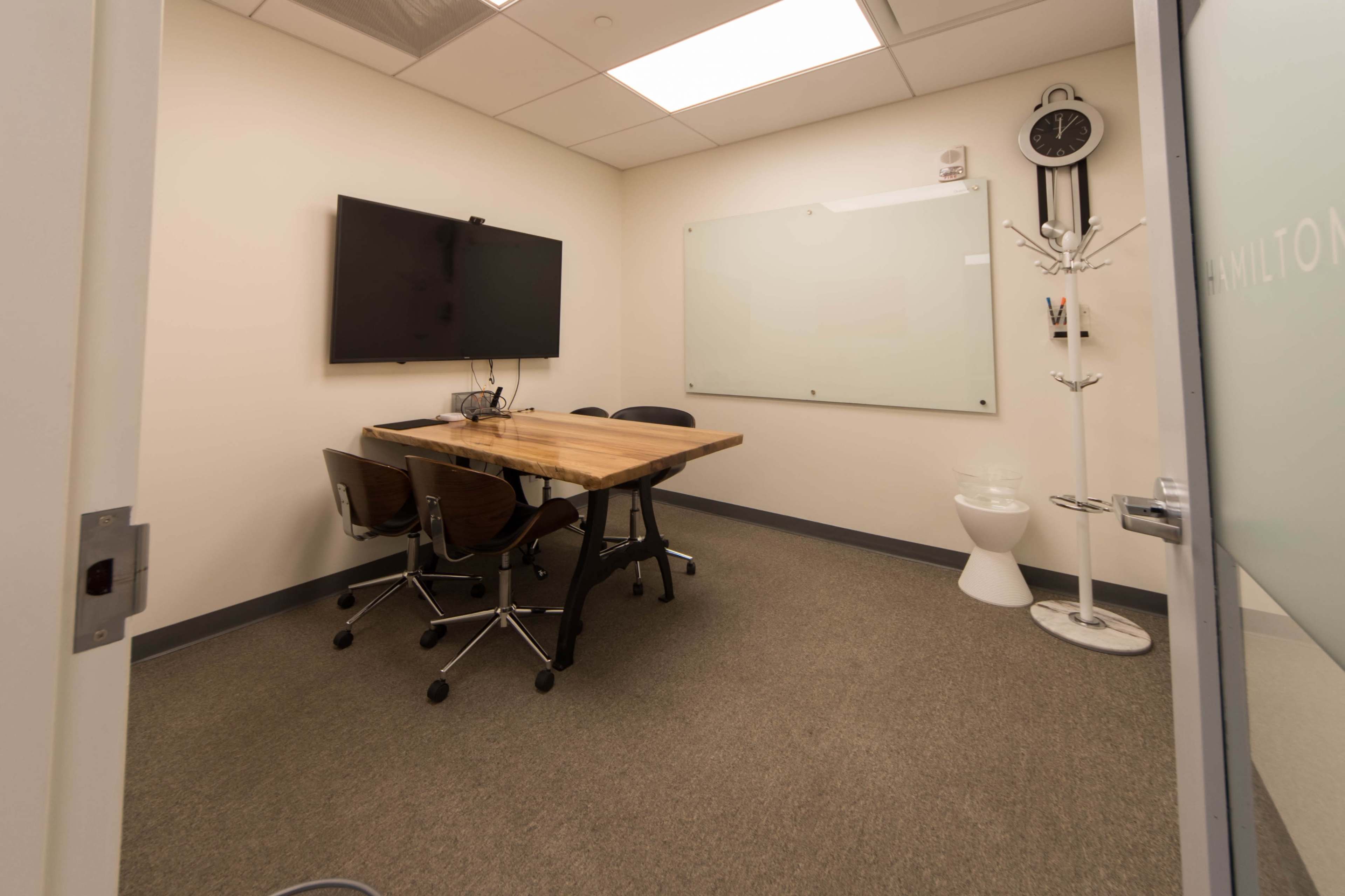 The image shows a small meeting room with a wooden table, two chairs, a wall-mounted screen, a whiteboard, and a coat stand.
