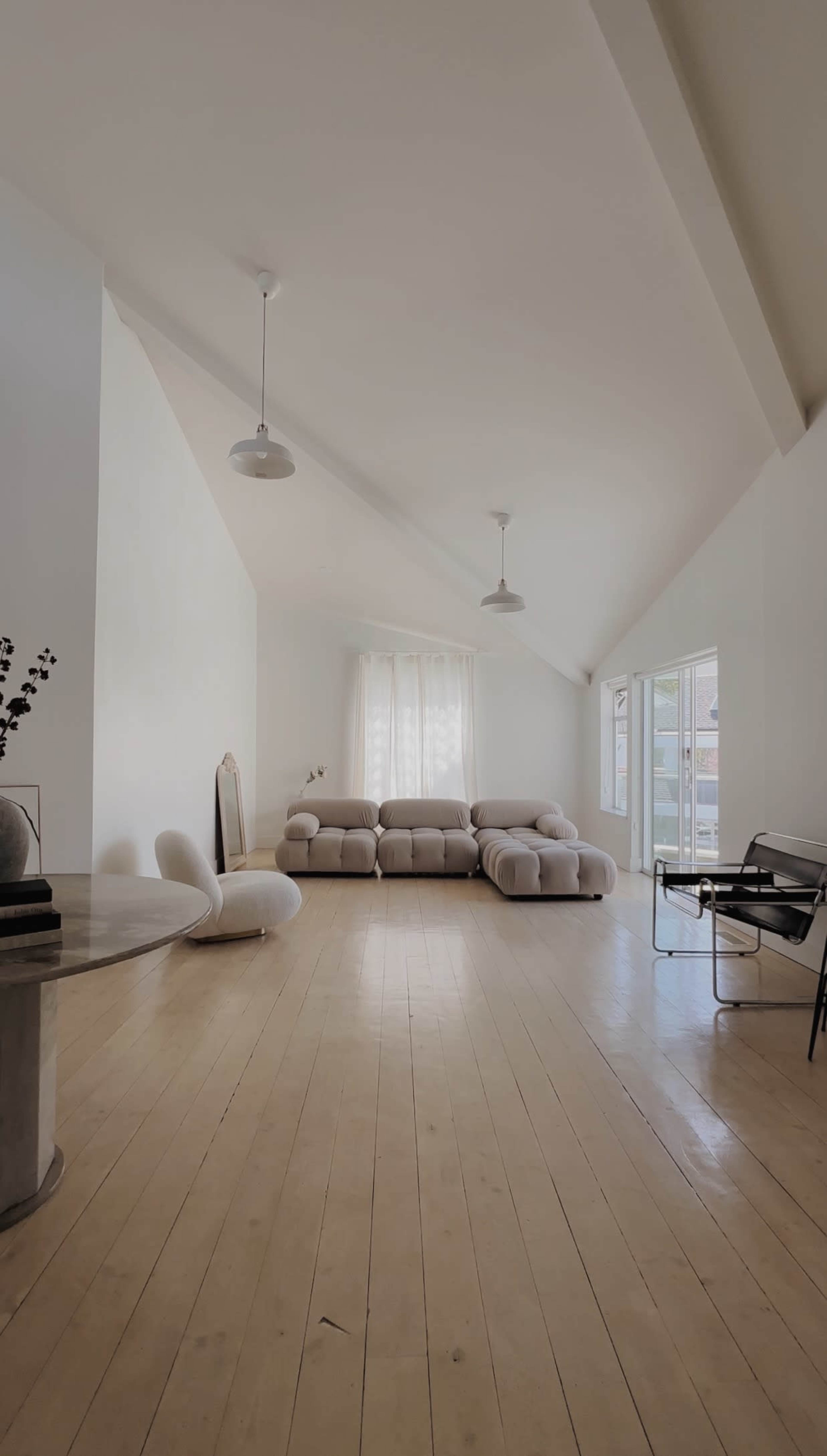 A minimalist living room with a light-colored sofa, a round table, and large windows allowing natural light to enter.