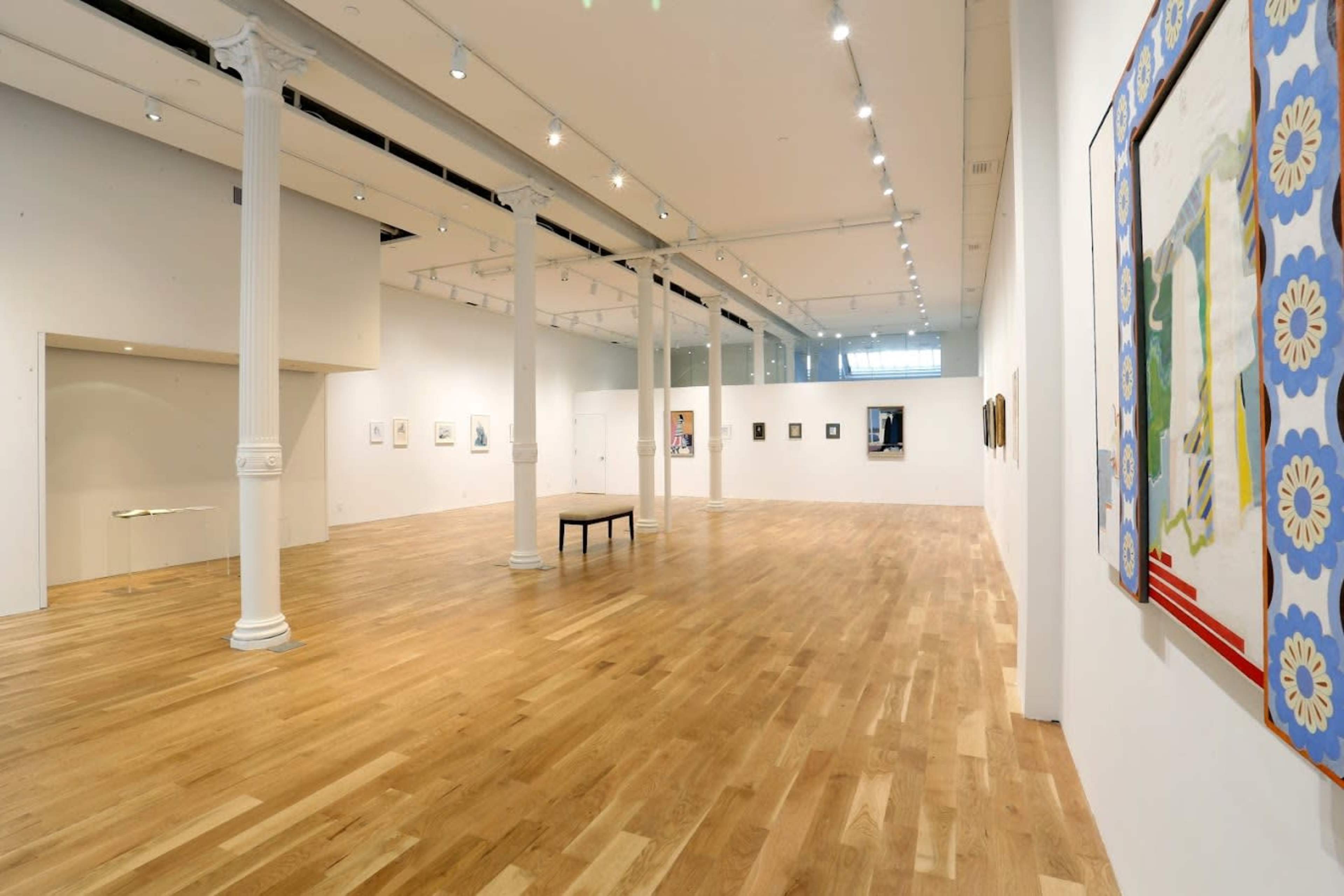 The image shows a modern art gallery with wooden flooring, white walls, and various artworks displayed along the walls.