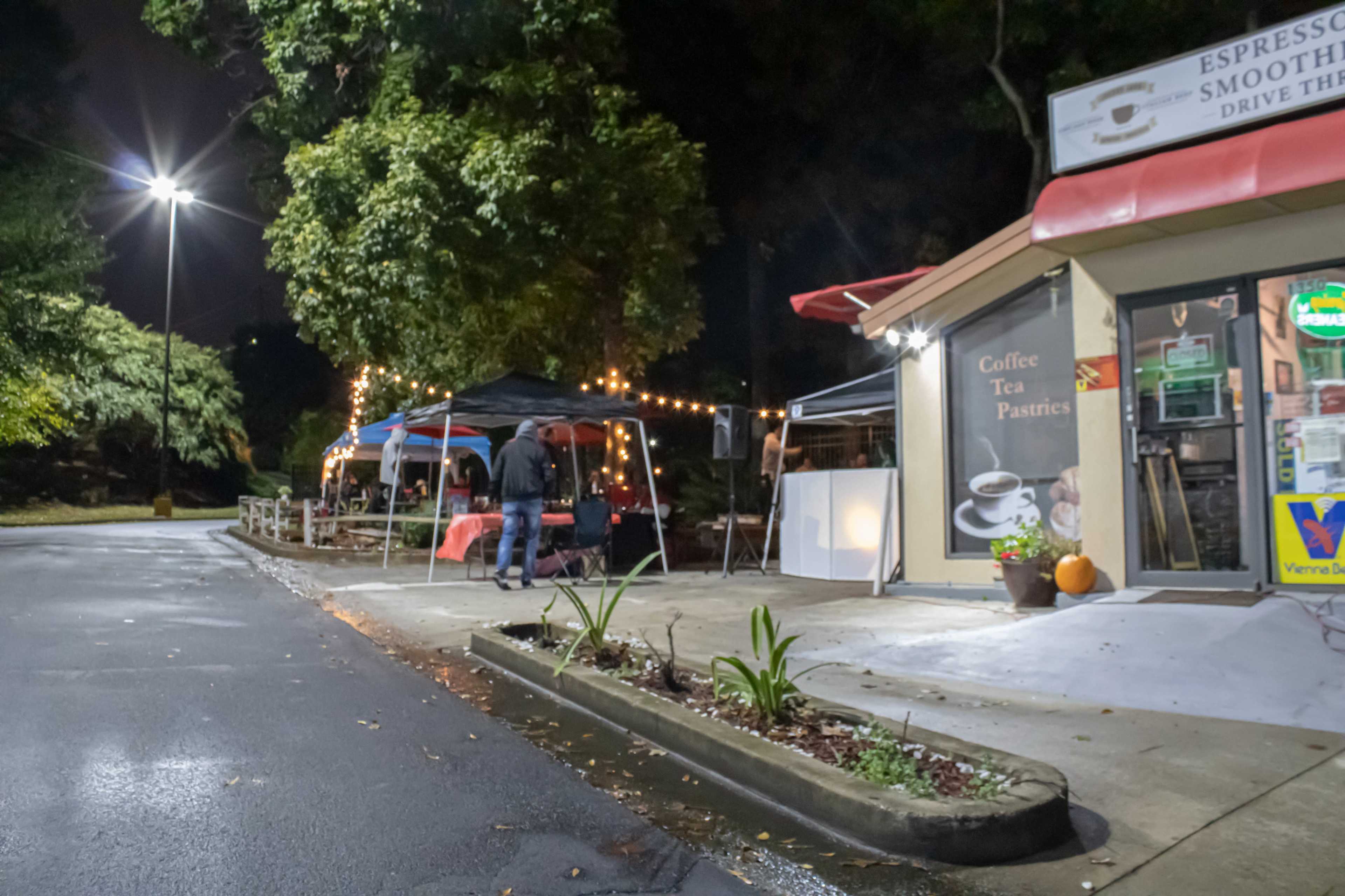 A small café with outdoor seating and string lights is set up on a quiet street at night, alongside a driver’s lane and a patch of greenery.